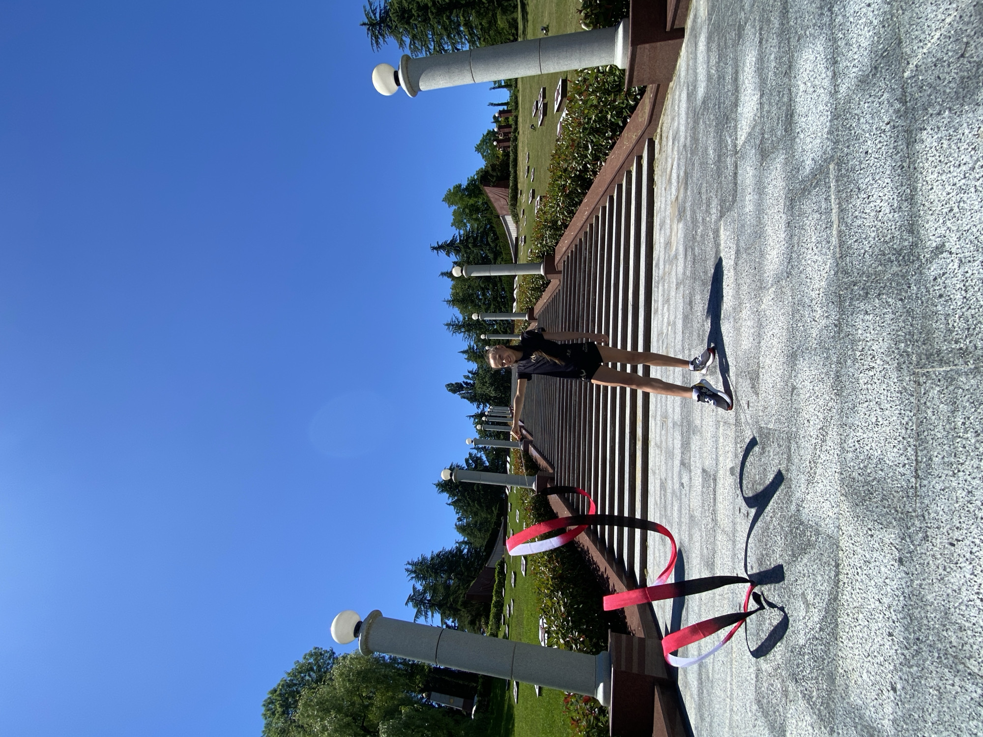 A young woman stands on a sunlit paved area, which appears to be part of a memorial or monument site. She is wearing a black t-shirt, black shorts, and athletic shoes. Her arms are outstretched to her sides, and she is looking at the camera with a slight smile, suggesting a relaxed and happy mood.
The setting is outdoors, under a clear, bright blue sky, indicating a sunny day, likely midday given the strong shadows. The paved area has a speckled granite texture and is bordered by a dark railing. Beyond the railing, there is a series of steps leading down to a grassy area with several tombstones or markers, characteristic of a cemetery or memorial park. Tall trees and shrubs surround the area, adding to the serene atmosphere.
There are decorative elements present, including tall, cylindrical pillars with spherical lights on top, placed at intervals along the paved walkway. Additionally, a red, white, and black ribbon is draped over the railing in the foreground, adding a splash of color to the scene. The overall impression is of a peaceful and well-maintained outdoor space. The presence of tombstones and the overall solemn design of the memorial suggest this could be a place of remembrance. No visible text is present in the image.