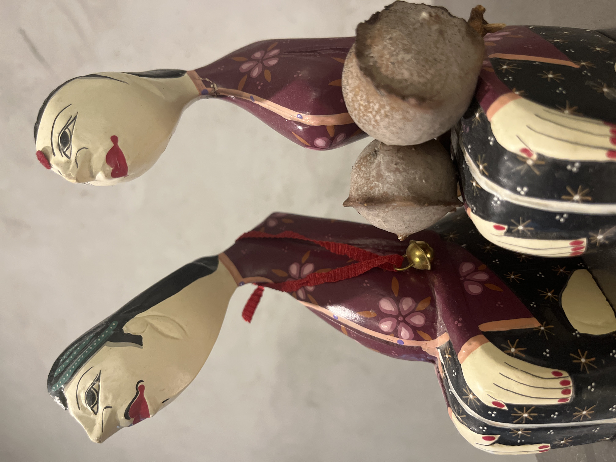 The image shows two wooden figures of women, lying on their sides against a pale background. The figures have light skin, black hair, and red markings on their faces. They wear clothing with floral patterns in shades of purple, pink, and brown. A red ribbon with a small brass bell is attached to one of the figures. There are two spherical objects, possibly seed pods, positioned between the figures. There is no information to indicate the location of the image being taken in Budva, Montenegro.