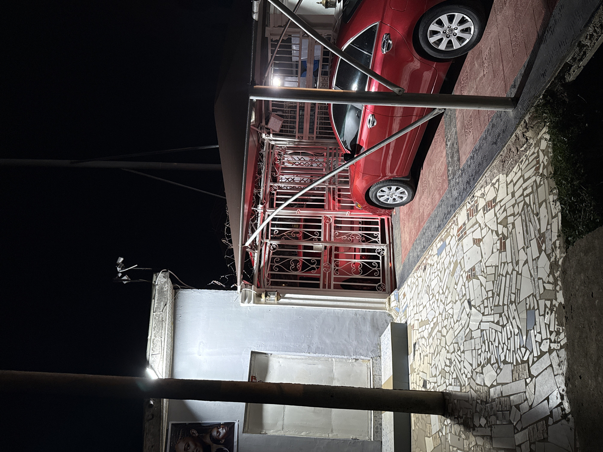 The image depicts a nighttime scene in Abuja, Nigeria, featuring a red sedan parked under a dark canopy. To the left, a light-colored building with two doors is visible, adjacent to a large, dark pole. The building has a poster with images of faces on its left side. A decorative, ornate metal gate, illuminated by red light, stands between the building and the car. The ground is covered in a mosaic of broken tiles and red paving stones. Several security lights and cameras are mounted on the building and near the gate.