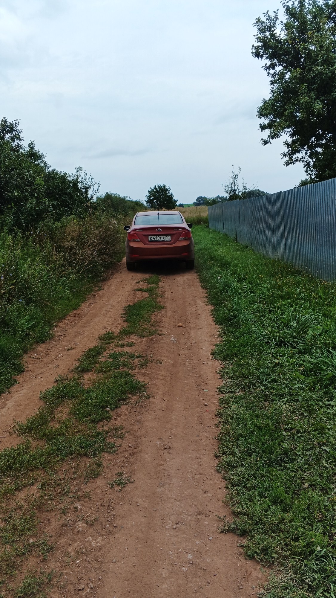 A red sedan is parked on a dirt road in the village of Podgornoye, Russia. The car, a Hyundai Solaris, is facing away from the camera and is parked on the right side of the road. The road is unpaved and has tire tracks from vehicles. There is grass growing on the sides of the road. A corrugated metal fence is visible to the right of the road, separating it from a field. Trees and bushes are present on the left side of the road, and in the background. The sky is overcast. The license plate of the car is visible and reads K419PX 18.