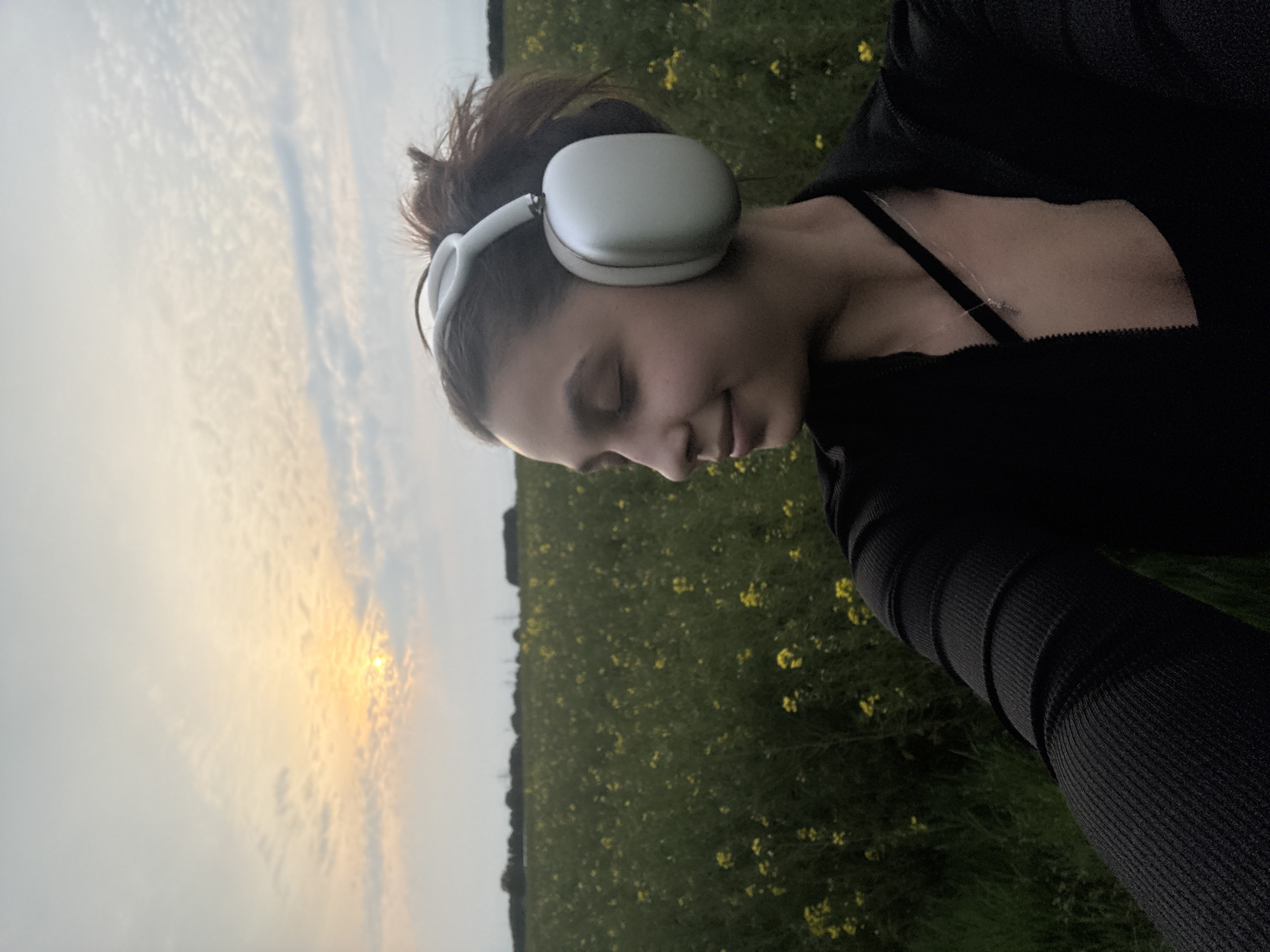 A person wearing white headphones and a black top is standing in a field of yellow flowers. The person has their eyes closed and appears to be at peace. In the background, there is a sunset. The city of Mönchengladbach, Germany is not visibly discernible in this image.
