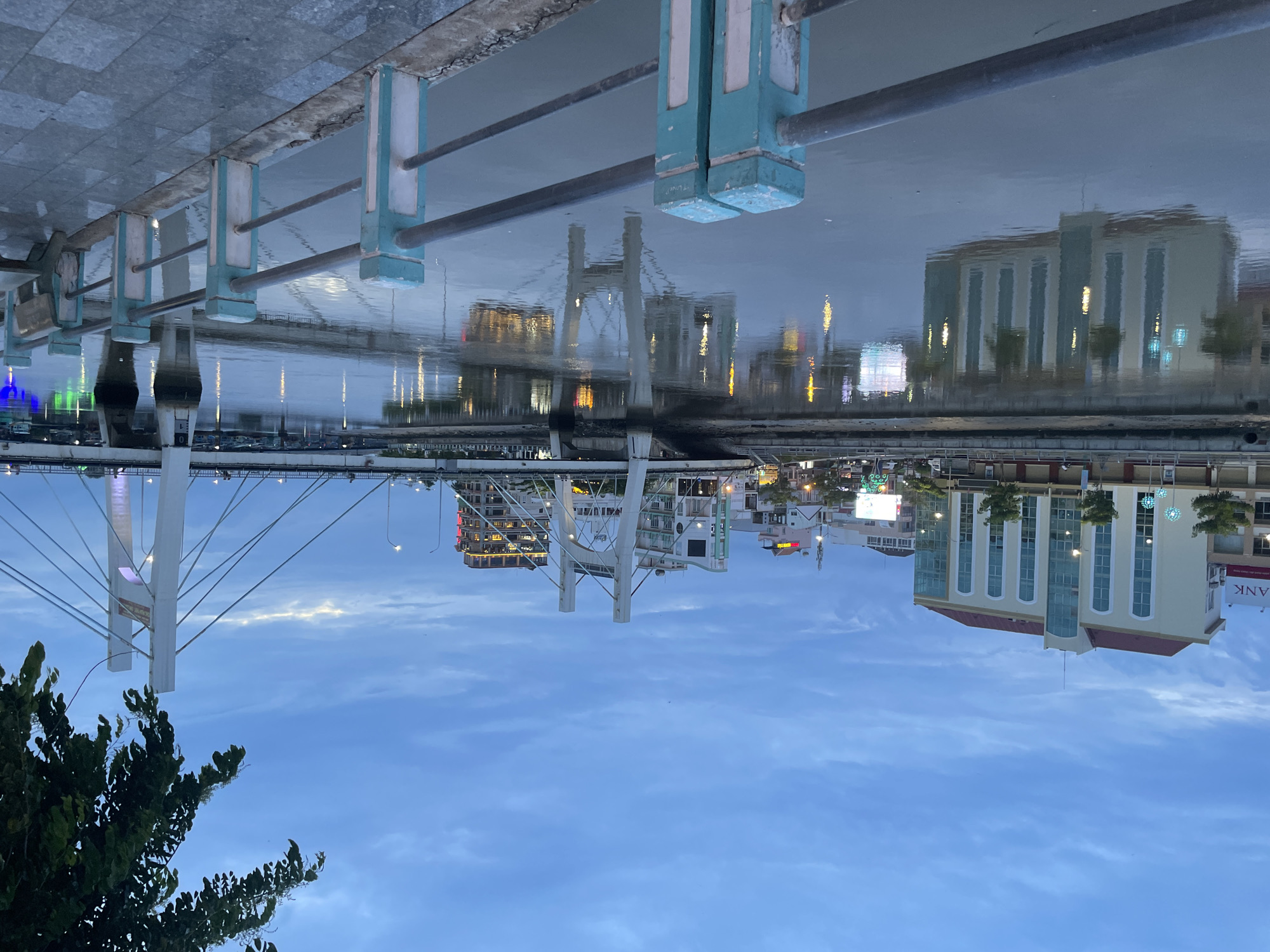 The image captures an urban waterfront scene at dusk or early evening, rotated 90 degrees clockwise from its natural orientation. When viewed correctly, a concrete promenade with a light blue and white painted railing and dark metal handrails runs along the left side. This railing separates the viewer from a calm body of water that occupies the center of the frame, extensively reflecting the sky, city lights, and structures.
Across the water, various buildings are visible, some illuminated with golden lights, which create vibrant reflections on the water's surface. A distinctive white bridge, possibly a cable-stayed or suspension type, spans the water with prominent white pylons and numerous support cables. The bridge structure is also clearly reflected in the water below it.
To the far right, a light-colored building with large vertical windows and a red-tiled roof stands out. Partially visible on its side, in white letters on a red background, is the word "BANK". Another building with illuminated windows can be seen further left along the bank. The sky above is a deep blue with scattered, light clouds, indicating either twilight or pre-dawn conditions. A lush green tree or bush is visible in the lower foreground (left when correctly oriented). The overall atmosphere is tranquil and serene, highlighting the interplay of light and reflections on the water. Given the provided context, the scene is located in Phan Thiết, Vietnam.
