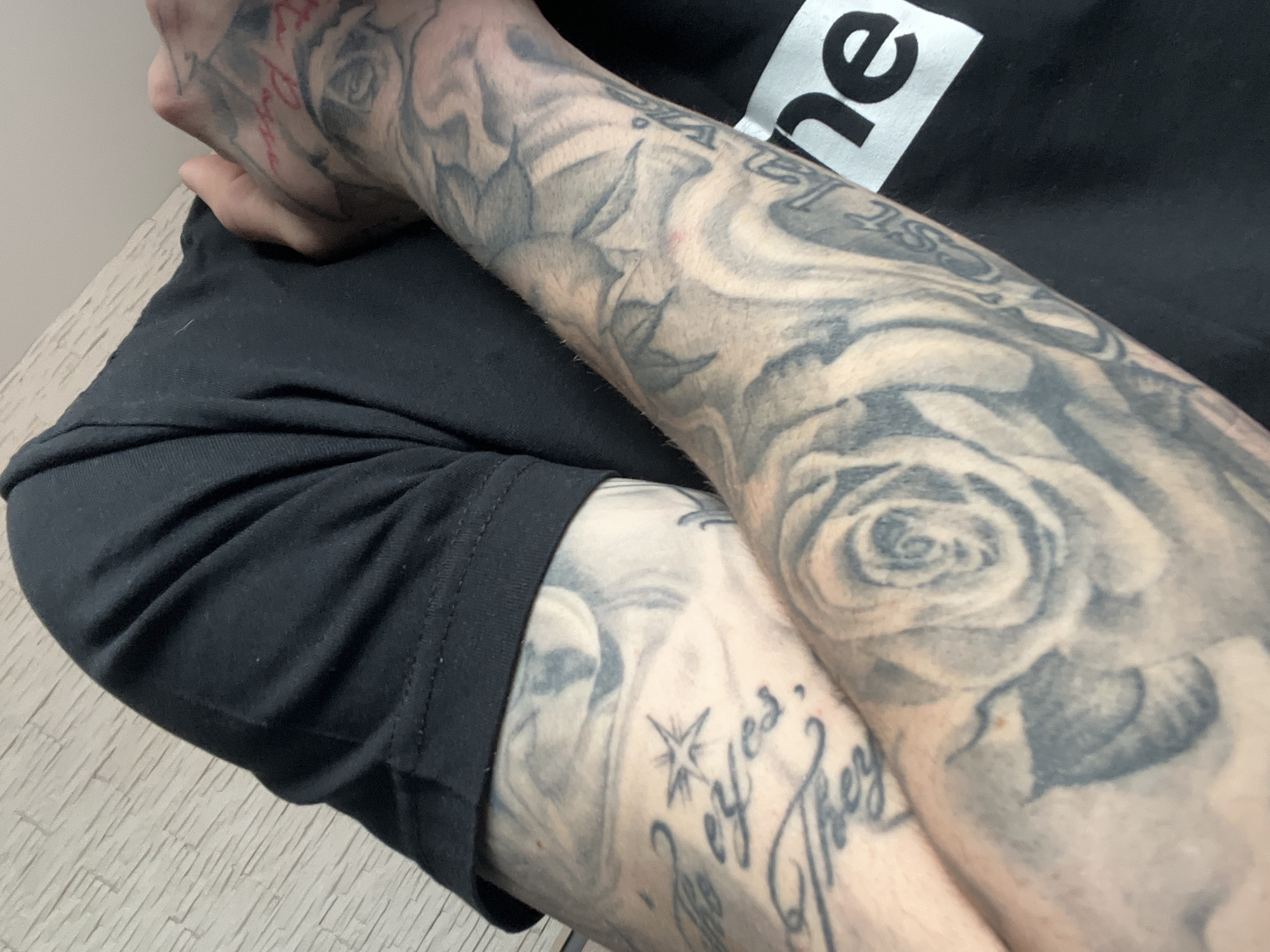 The image shows a person's arms and part of their torso. The person is wearing a black t-shirt with a white box logo containing the letters "ne". The arms are heavily tattooed with black and grey designs, including roses, flowers, and lettering. The person is sitting down, with one arm resting on the other. The background is blurry, but suggests a simple interior space. There is no indication of the image being taken in Belovo, Bulgaria.