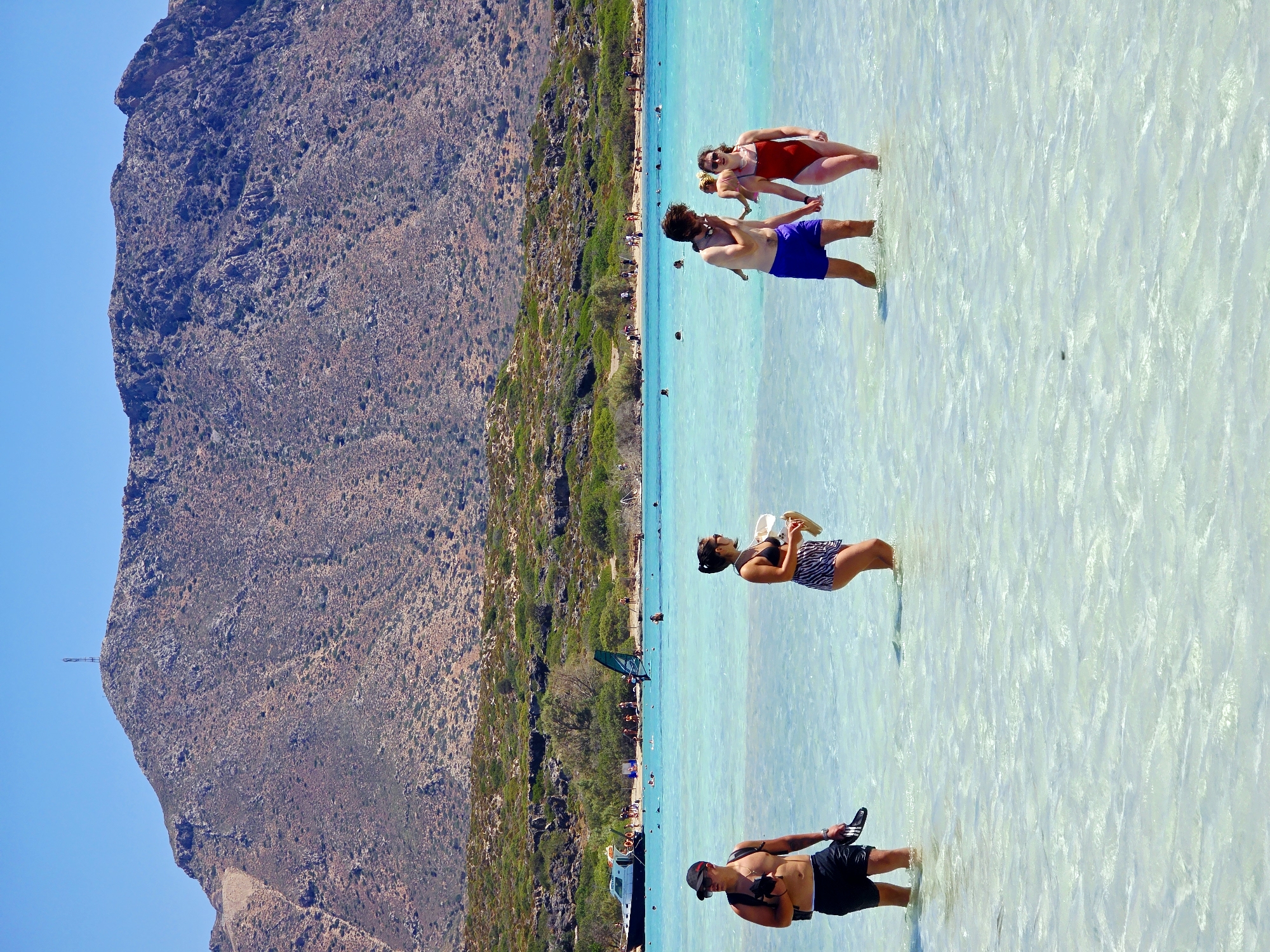 Four people are wading in shallow, clear turquoise water on a sunny day. In the foreground, on the left, a man wearing black swim trunks and a black baseball cap is holding a pair of black flip-flops and a dark object, possibly a camera. To his right, a woman in a black bikini top and black and white striped skirt is holding a white object, possibly a towel, and a pair of light-colored sandals. Further to the right, a couple is walking hand-in-hand. The man is wearing blue swim trunks and has a snorkel mask around his neck. The woman is wearing a red swimsuit. The water is shallow enough that their legs are visible.
In the middle ground, the turquoise water extends to a beach lined with green shrubs and some trees. A white boat with black trim is moored near the shore, and a person on a windsurfing board is visible. Several other people are scattered on the beach and in the water.
The background features a large, rocky mountain under a clear blue sky. The mountain is covered in sparse vegetation and has a dark, conical structure at its summit. The overall scene depicts a lively beach environment.