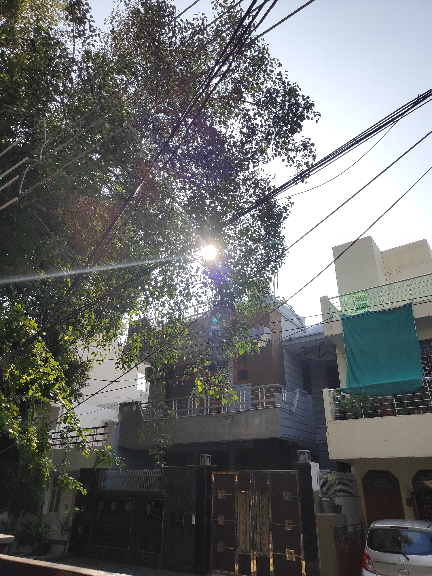 The image captures a street view in Gurgaon, India, focusing on residential buildings and overhead utility lines. A large tree with dense green foliage is prominent in the foreground, its leaves partially obscuring the view of the sky and the buildings behind. Sunlight streams through the branches, creating lens flare and glinting off the utility wires that crisscross the scene.
Behind the tree and wires, a multi-story residential building is visible. The building features a modern architectural style with balconies, a grey and white facade, and decorative elements. One balcony on an upper floor is draped with a large, dark green tarp. Below the upper structure, a darker-colored, ornate metal gate is visible, suggesting an entrance to a property. The gate appears to be closed.
To the right of the main building, another section of a residential structure is present, with a cream-colored upper facade and a balcony. The bottom portion of this structure shows a door and a window with a barred grille. A silver car is parked to the right, partially visible in the lower right corner of the frame. The sky above is clear and bright, indicating daylight conditions. The scene depicts a typical urban residential environment in India, characterized by dense foliage, overhead wiring, and multi-story homes.