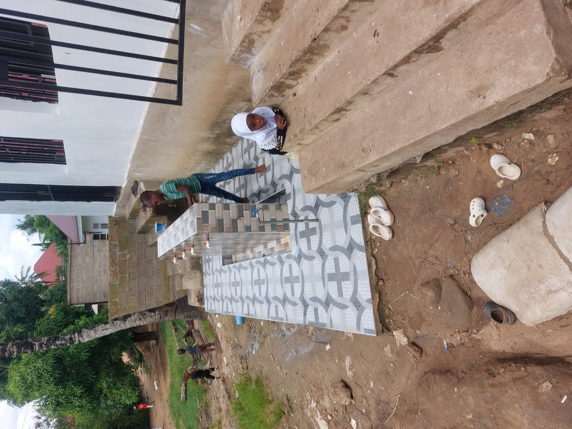 Two children are present: a young girl wearing a white hijab and a young boy washing his hands at an outdoor washing station. The washing station consists of multiple faucets mounted on a tiled structure. The surrounding environment is dusty with scattered debris and some discarded shoes. In the background, there is a two-story building with black-framed windows. In the distance, tropical trees, a red roof, and other buildings are visible. Given the image's origin and visual cues, it is likely in Dar es-Salaam, Tanzania. Other people walk behind the washing station, near trees.