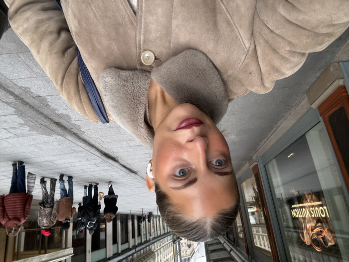 A selfie captures a woman in a tan shearling coat, wearing a white earbud. She stands on a stone-tiled sidewalk in front of a storefront, reflecting the iconic Louis Vuitton logo. Several people walk behind her along the sidewalk. The surrounding architecture features traditional buildings, hinting at an urban European setting. The image's perspective is slightly tilted.