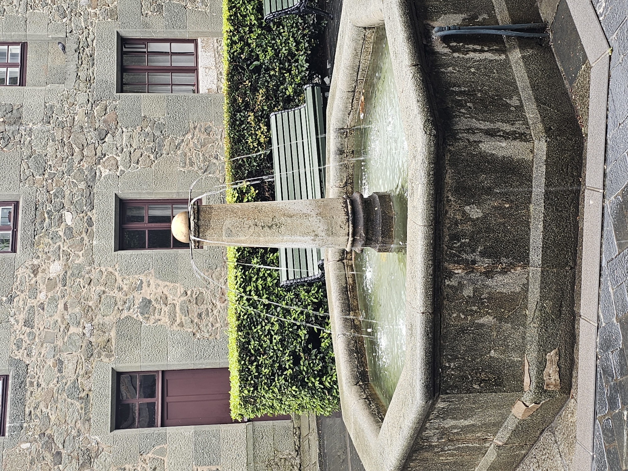 The image shows a stone fountain in Алупкинский городской совет, Ukraine, set against a building with stone walls and windows with dark frames. A green hedge and a green bench are located behind the fountain. Water streams from the central pillar of the fountain into a stone basin filled with water. The fountain's structure shows signs of weathering and minor damage. The ground around the fountain is paved with stone tiles.