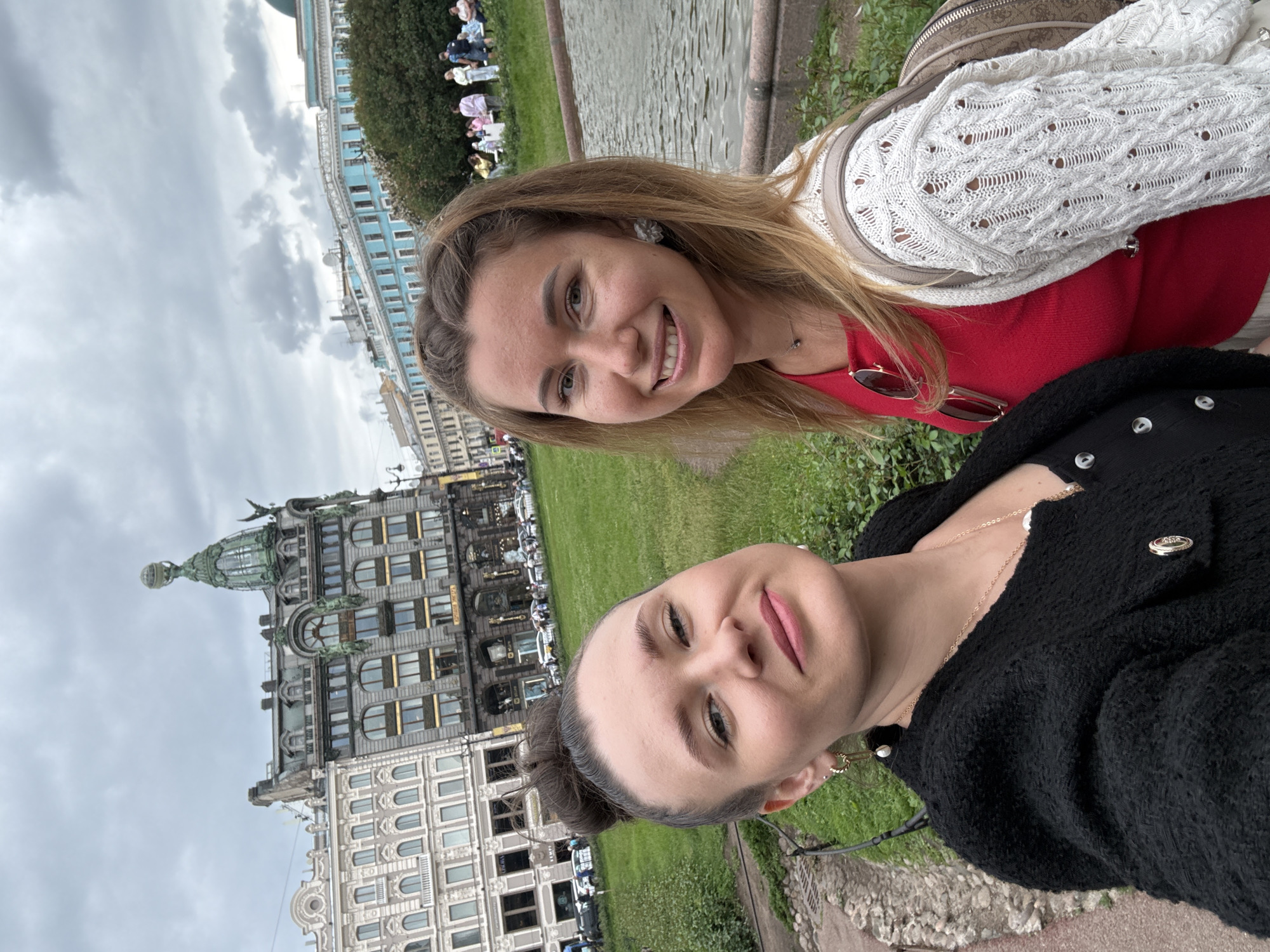 Two young women are taking a selfie outdoors in Saint Petersburg, Russia. The woman on the left has her hair pulled up in a bun and is wearing a black textured jacket with gold buttons. She has on gold hoop earrings with pearls and a gold necklace. The woman on the right has blonde hair and is wearing a red top and a white crocheted cardigan. She is also wearing a floral earring and a small necklace. Behind them, there are buildings, a grassy area, and a body of water. The building in the center background is distinctive with a green dome topped with a globe. This is the Singer House, also known as the Dom Knigi (House of Books), located on Nevsky Prospekt in Saint Petersburg. The sky is overcast.