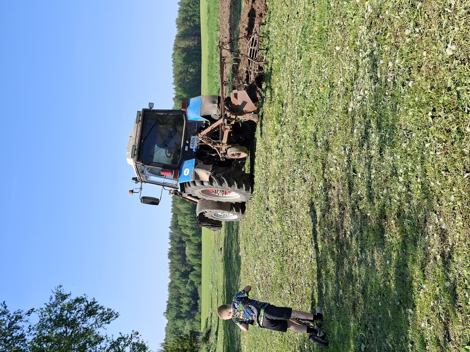 A child in a patterned shirt and black shorts lies on a grassy field, casting a long shadow. A blue and white tractor with an attached tiller is parked on the edge of a tilled field. A forest is in the background under a clear blue sky. The license plate on the tractor reads "4806 SC 58". The scene is in a rural setting. There is no indication in the image that the scene is in the city of Karsha, Russia.