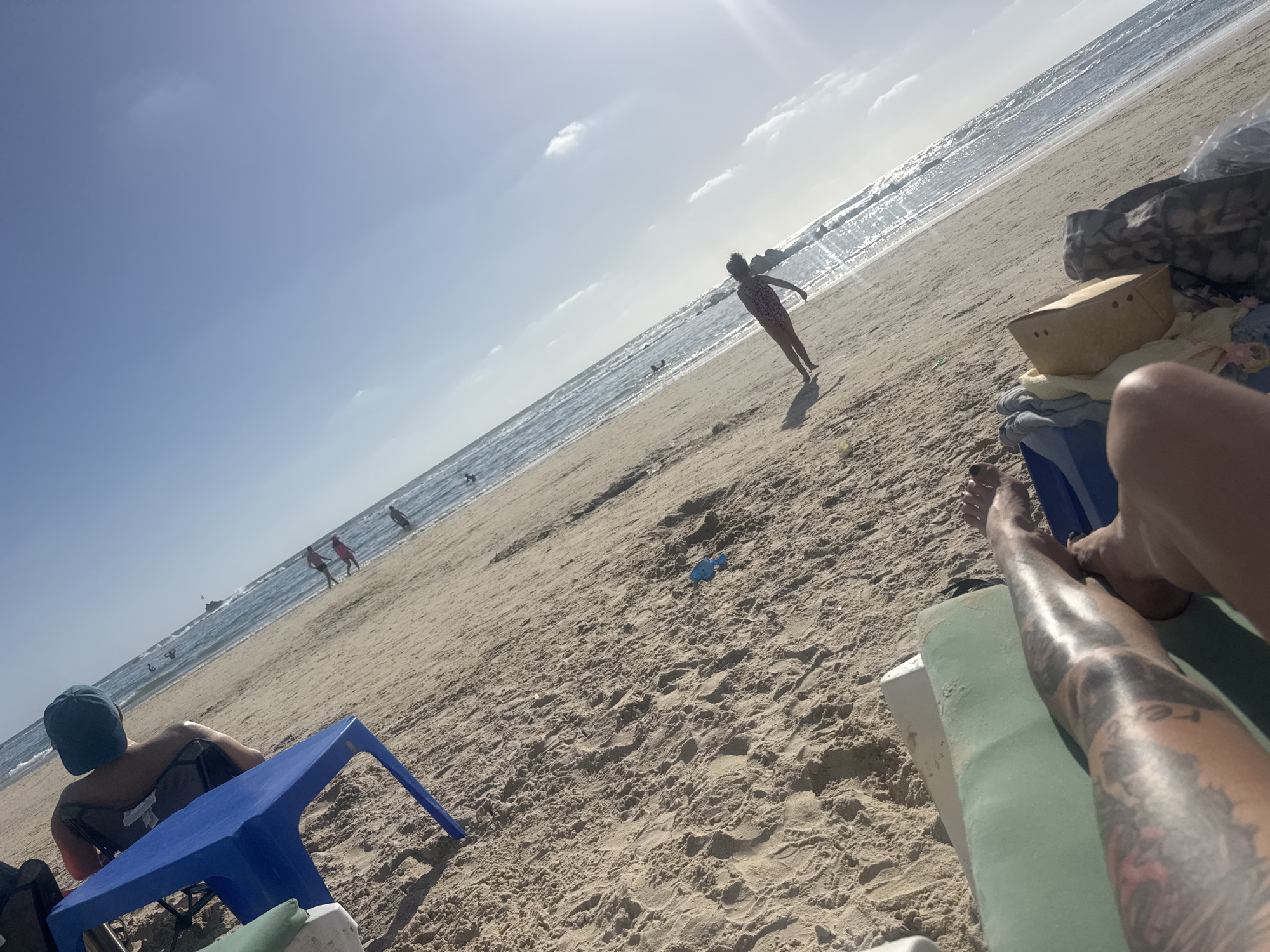 The photograph was taken on a beach in Bat Yam, Israel, on a sunny day. The beach has light-colored sand. In the foreground, a person's leg with tattoos rests on a green and white beach lounger. Another person sits in a blue plastic chair, facing away from the camera. Further along the beach, a person in a swimsuit stands facing the sea, with arms outstretched. Other people can be seen walking or swimming in the distance. A blue plastic toy lies on the sand. Beach gear and belongings are piled to the right of the lounger. The sea is visible in the background, with sunlight reflecting on the water. The sky is light blue with some clouds.