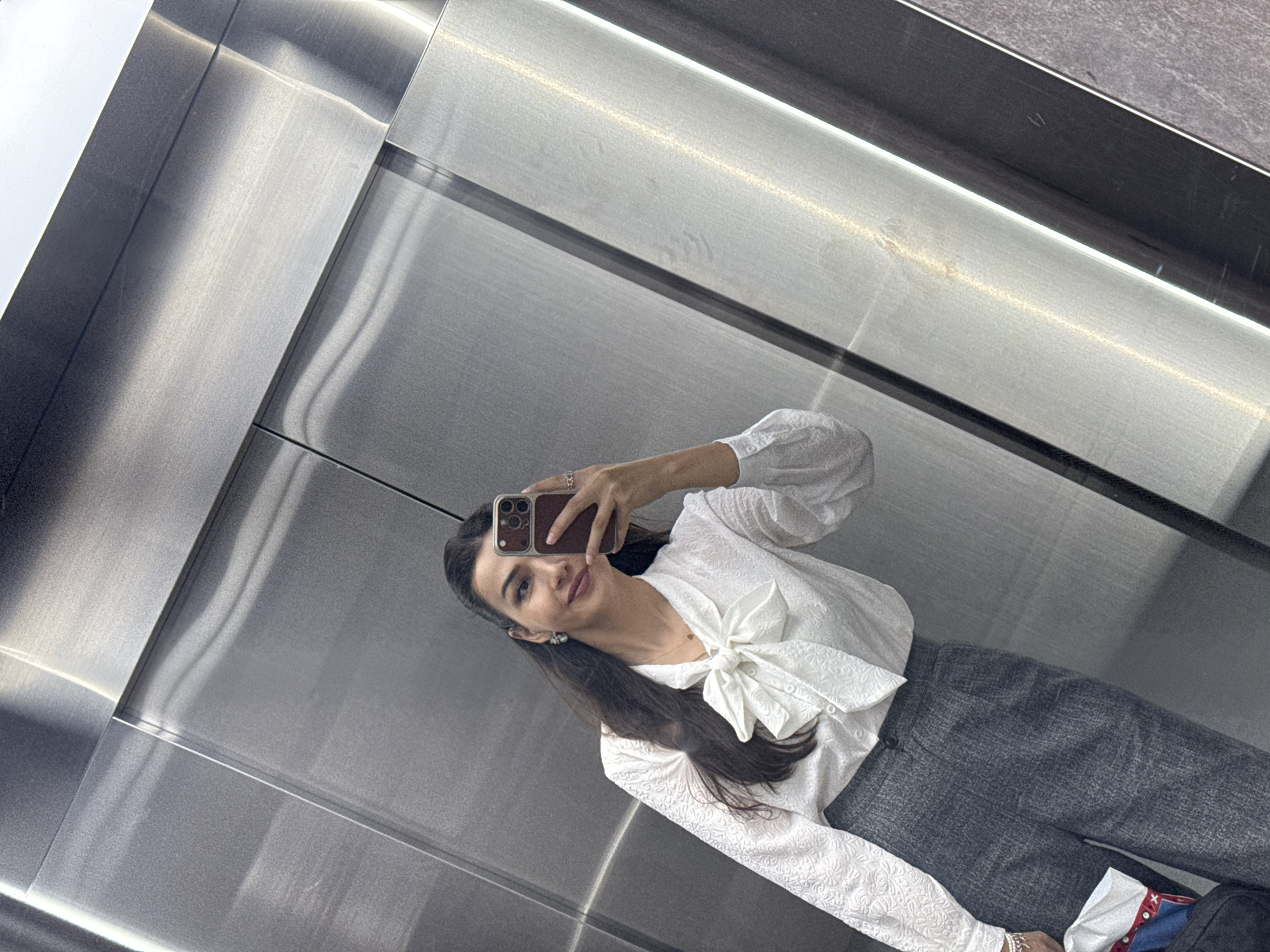 A woman is taking a mirror selfie in an elevator. She is wearing a white textured blouse with a large bow at the neck and grey patterned pants. She has long dark hair and is wearing earrings and a necklace. She is holding a phone in her right hand to take the photo. The elevator walls appear to be brushed metal. The location is Sabunchu Raion, Azerbaijan.
