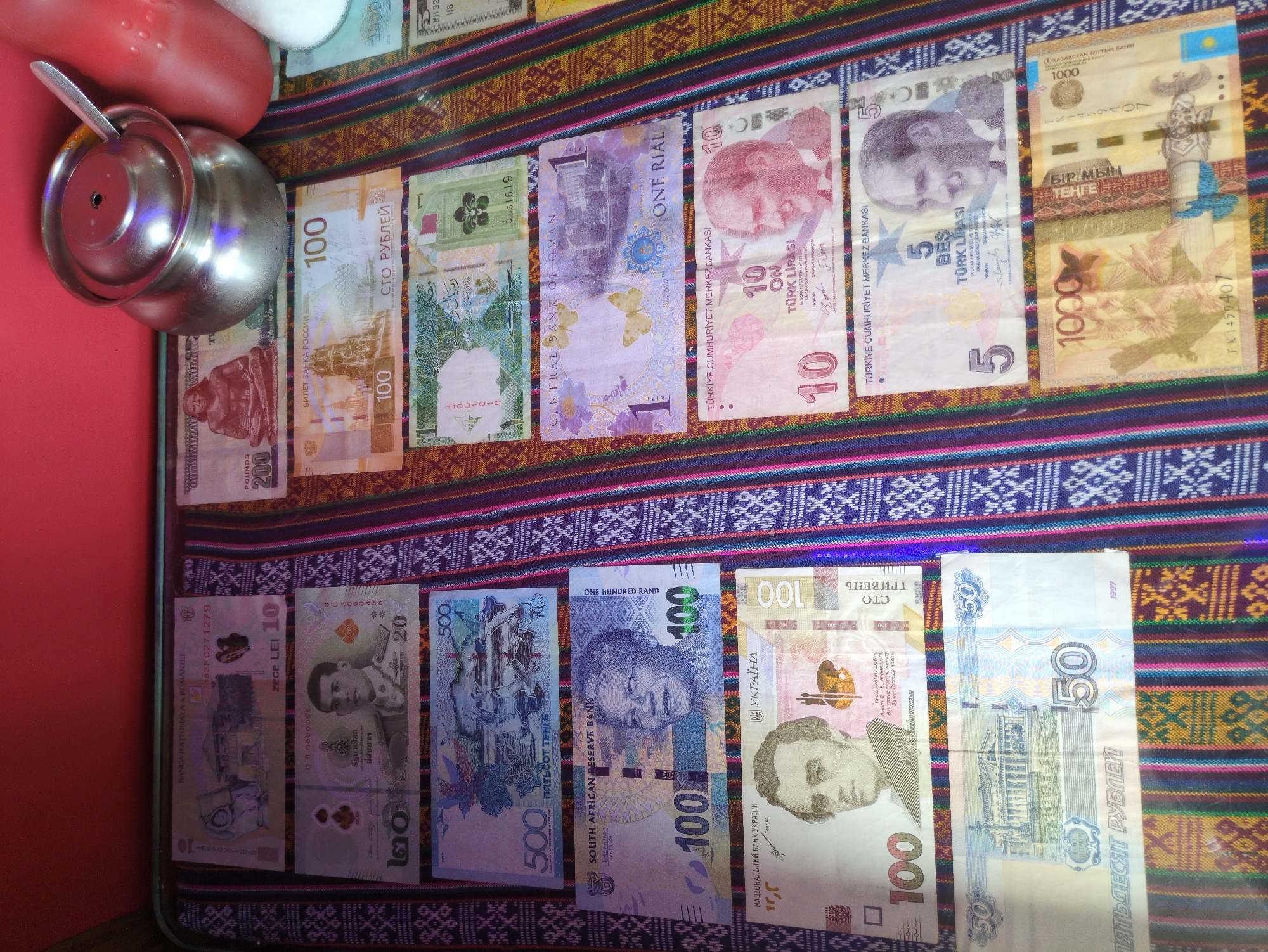 The image displays a collection of various banknotes laid out on a colorful, striped textile surface. The banknotes appear to be from different countries and denominations. Some of the visible currencies include Romanian Leu, Thai Baht, Russian Ruble, South African Rand, Ukrainian Hryvnia, and Omani Rial, among others. A metal pot with a lid and a spoon rests on the top right of the textile. The background is out of focus, but hints at a red surface and a white, translucent object. The scene is a still life arrangement of currency and household items, suggesting it may be a personal display or collection.