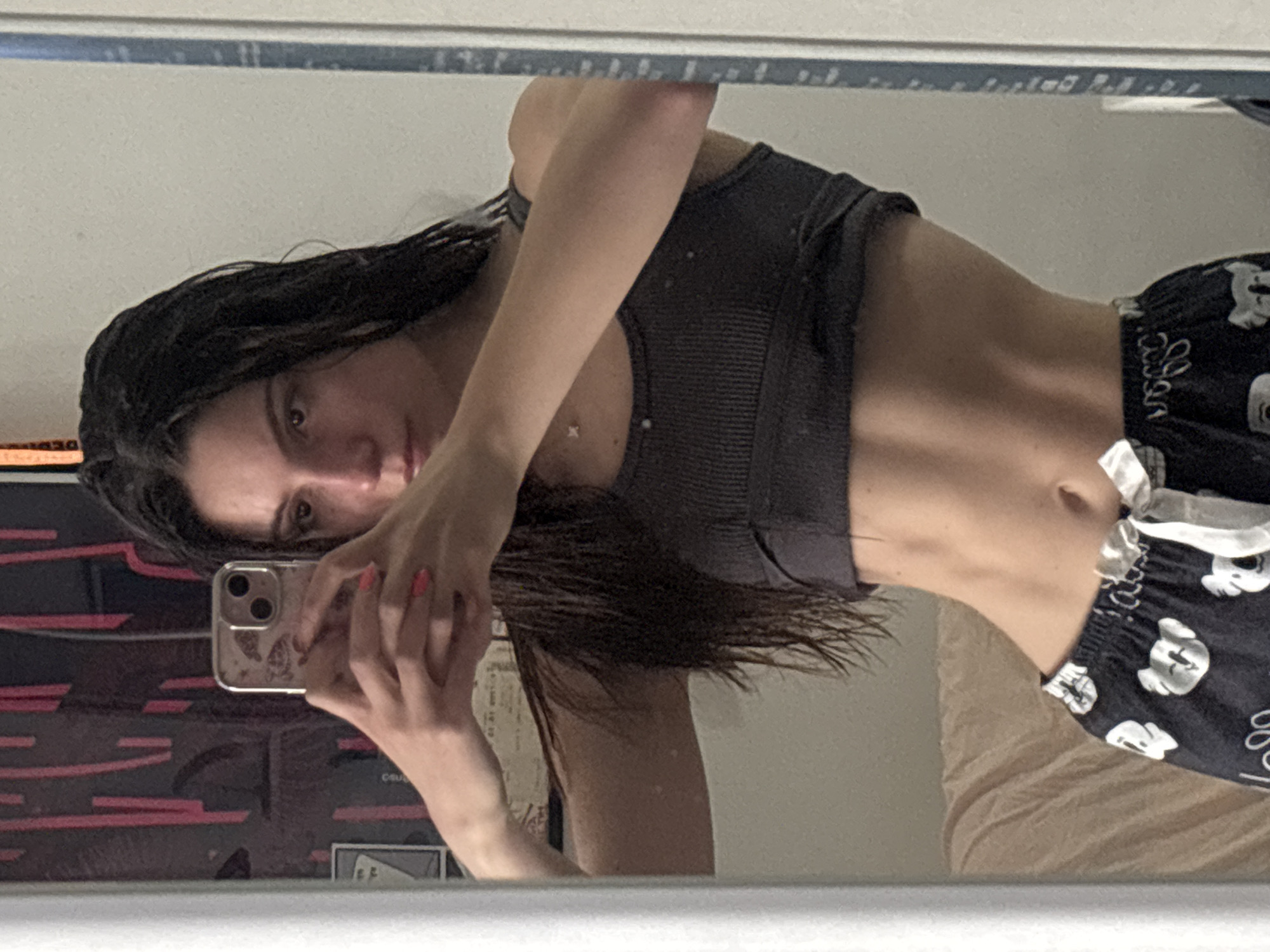 A person is taking a selfie in a mirror. The person has long dark hair and is wearing a gray ribbed tank top and patterned pajama bottoms with a white drawstring. The person's phone is visible in their hand. The background is blurry, but there appears to be a poster and a beige-colored object in the mirror's reflection.