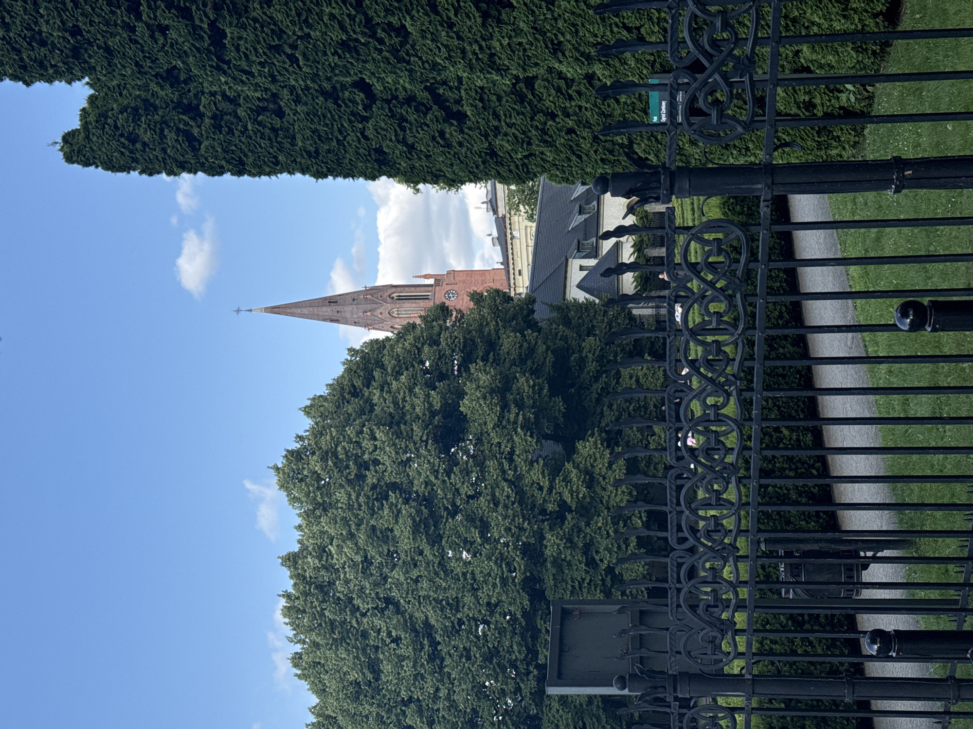 The image shows a view through a black wrought-iron fence towards a park area and buildings beyond. In the foreground, the ornate fence features curved scrollwork. Behind the fence, a paved pathway runs through a green lawn, bordered by manicured hedges. A black trash receptacle is visible behind the fence. In the middle ground, a building with a dark roof and dormer windows is partially obscured by trees. To the left, a large, dense green tree dominates the view. In the background, a tall, reddish-brown church spire with a cross on top rises against a blue sky with scattered white clouds. The architecture of the church and the park setting suggest it is located in a European city. Specifically, this scene captures the view of the Church of Saints Peter and Paul and the surrounding park in Poznan, Poland.