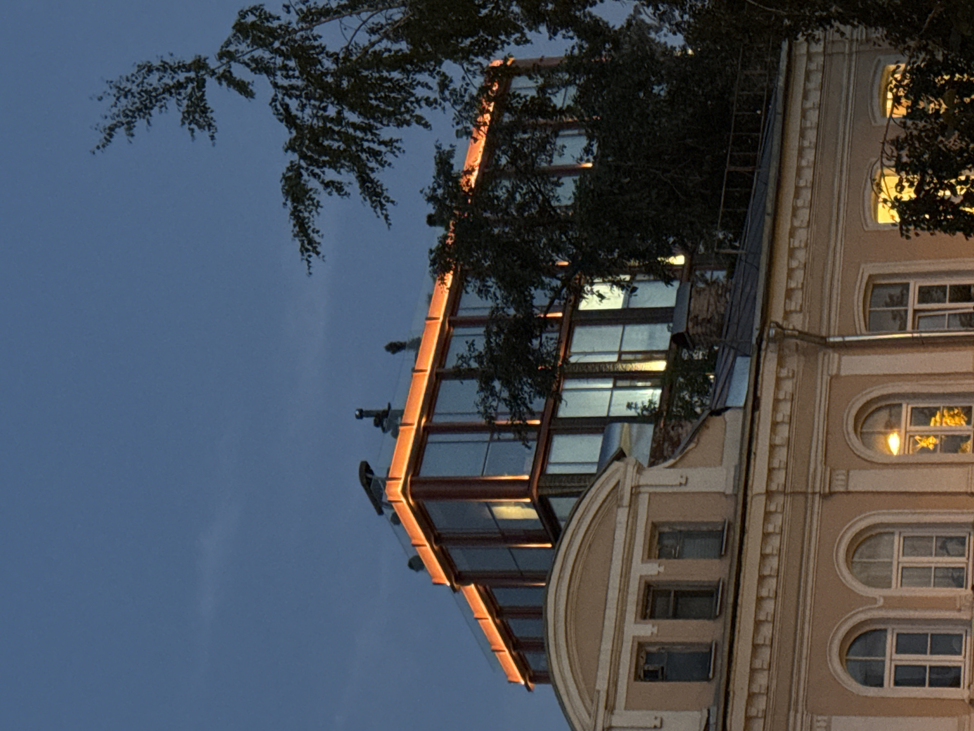The image shows a multi-story building with a modern glass-enclosed upper level illuminated by orange lighting. The building is situated in Moscow, Russia, specifically in the historic Arbat district. In the foreground, an older, light-colored building with arched windows and decorative molding is visible. Trees with dark foliage partially obscure the view of the modern structure. The sky is dark blue, suggesting it is dusk or nighttime. There are no people or discernible actions depicted in the scene.