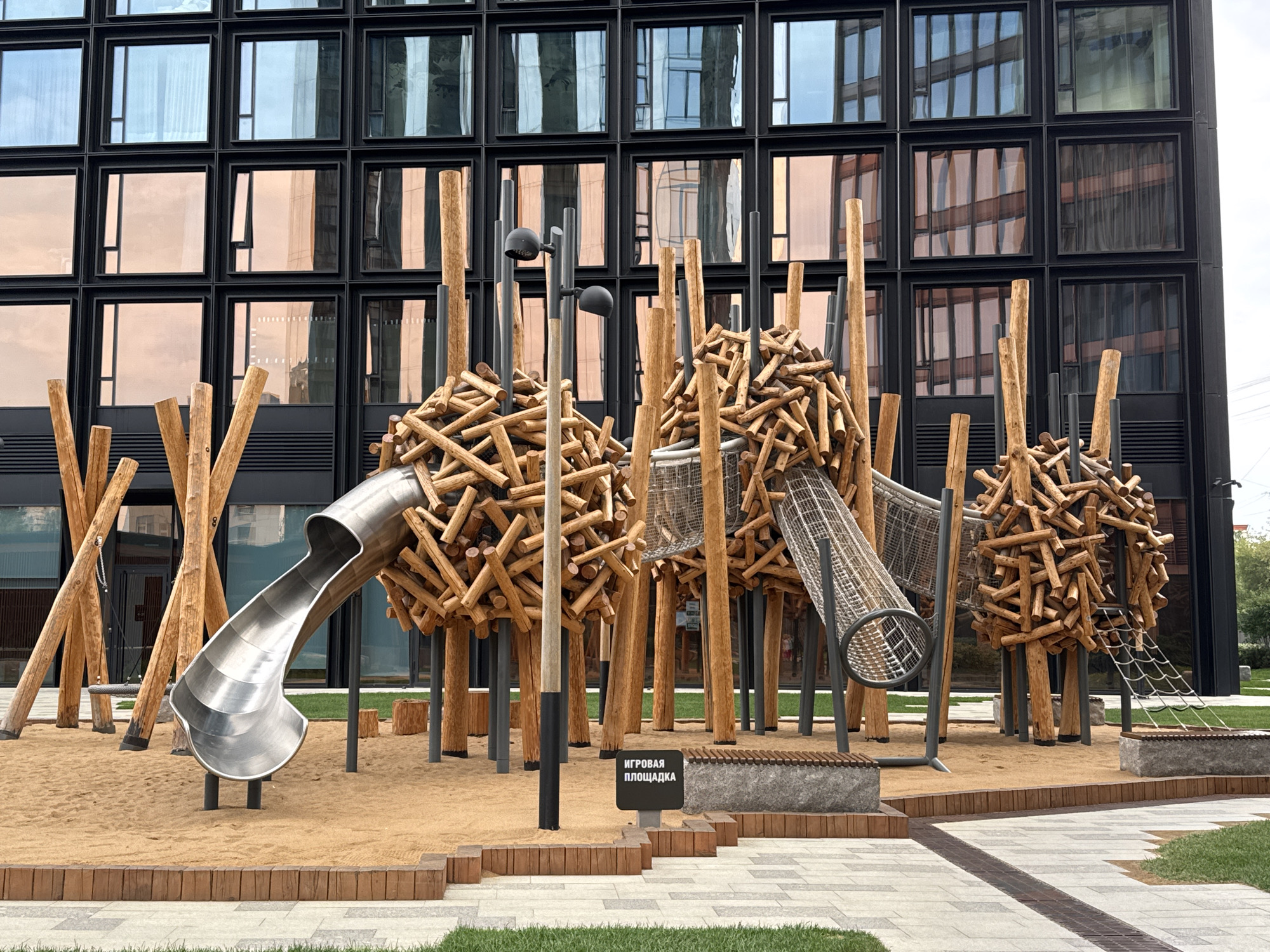 The image shows a modern playground constructed with natural materials, located in Khimki, Russia. The playground features large, spherical structures made of intertwined wooden logs, resembling nests. A silver, curved slide descends from one of these structures into a sandy area. Other elements include rope nets for climbing, wooden poles, and a metal grate. In the background, a contemporary building with a facade of dark frames and reflective windows dominates the scene. A sign with Cyrillic text, "ИГРОВАЯ ПЛОЩАДКА" (Playground), is visible in the sand. The ground is a mix of sand, paving stones, and grass. The overall atmosphere is clean and orderly, suggesting a newly developed urban space.