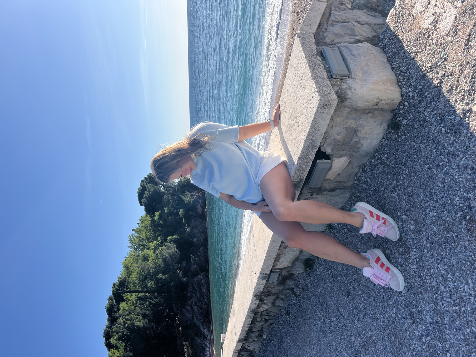 A young woman is captured in a bright, sunlit coastal scene, likely in Sveti Stefan, Montenegro. She is sitting or leaning on a low, light-colored concrete wall that runs parallel to the sea. The woman has long, light brown hair and is wearing a pale blue long-sleeved top, white shorts, and light-colored sneakers with distinct pink Adidas stripes and pink laces. Her gaze is directed downwards or to her side, and one hand rests on the concrete surface.
The setting features the calm, blue-green sea stretching into the distance, with gentle white waves breaking near the shore. Beyond the concrete wall and the water, a verdant hillside covered with dense green trees and foliage rises. In the immediate foreground, to the right of the wall, there is a gravel path or ground mixed with larger rocks, suggesting a natural and paved walkway.
The image conveys clear, sunny weather with a bright blue sky, where faint airplane contrails are visible. The shadows suggest it is mid-day or afternoon. The overall impression is one of a peaceful, scenic Mediterranean coastal environment.