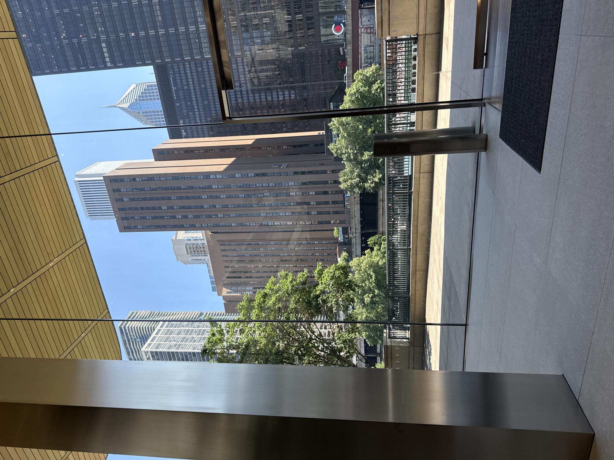 This is a horizontal, eye-level, outdoor photograph taken on a sunny day in Chicago, Illinois. The vantage point is from inside a modern building, looking out through large glass windows. In the foreground, a large, brushed metal pillar is on the left, and a glass door with a dark doormat in front of it is on the right. The floor inside is tiled. Through the glass, the Chicago River is visible, with trees and greenery along its banks. Several tall buildings of varying architectural styles form the cityscape beyond the river. One prominent building is made of brown brick with many windows, and another is a sleek, modern skyscraper with a distinctive pointed top. The sky is clear blue. There are no people visible in the image.