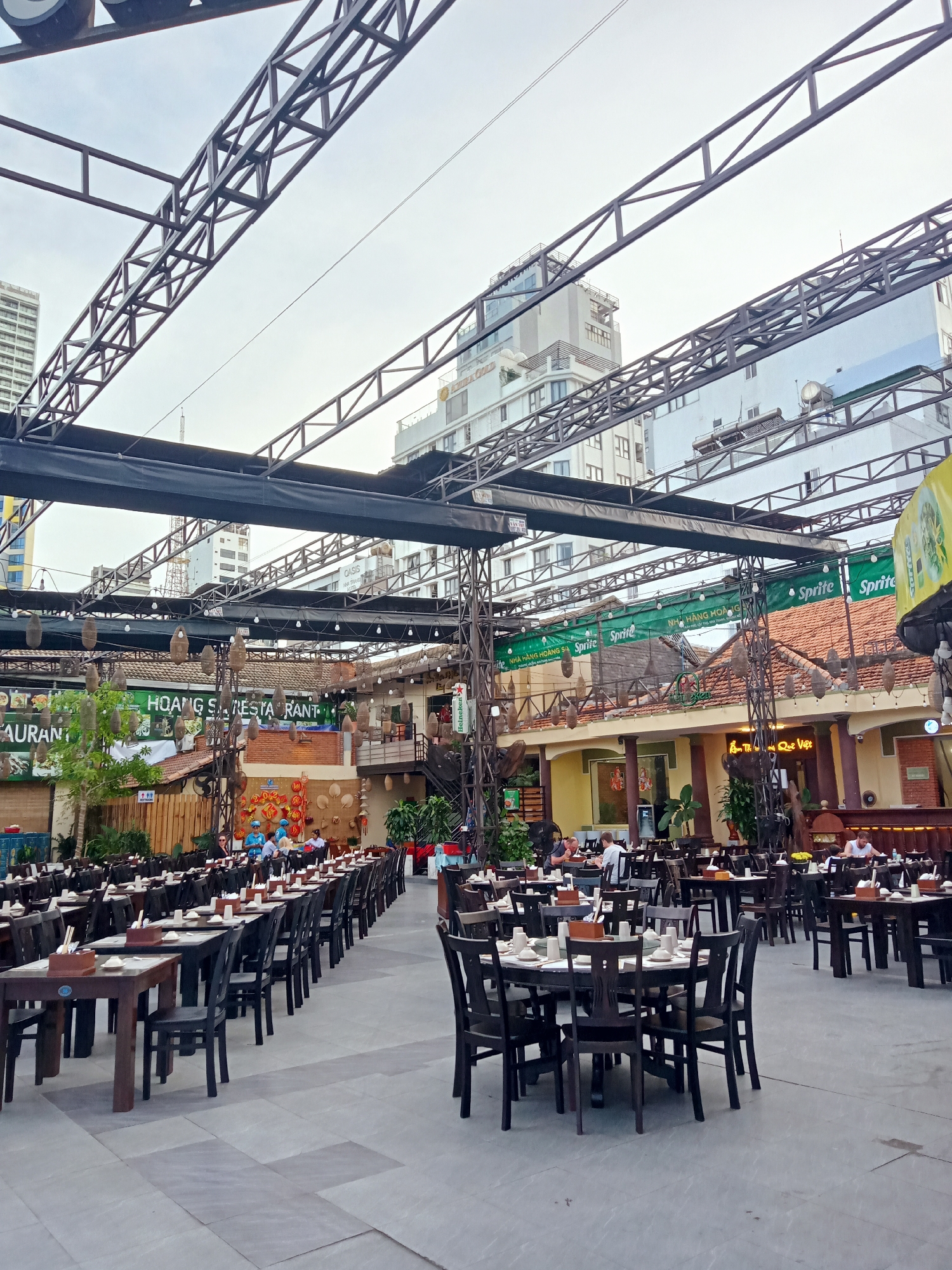 This image captures an outdoor dining area, potentially of a restaurant, in Nha Trang Ward, Vietnam. The space is densely populated with dark wooden tables and chairs, set up for dining. Patrons are seen seated at some tables, engaged in meals or conversations.
Overhead, a complex metal structure spans the area, possibly providing support for lighting or shading. Banners with 'Sprite' logos and the text "NHÀ HÀNG HOÀNG SA" are visible, indicating a specific restaurant or brand association. Additional signage features the name "HOANG SA RESTA RANT".
The background shows buildings of varying heights, typical of an urban environment. The overall scene suggests a busy, open-air dining experience in a commercial district.