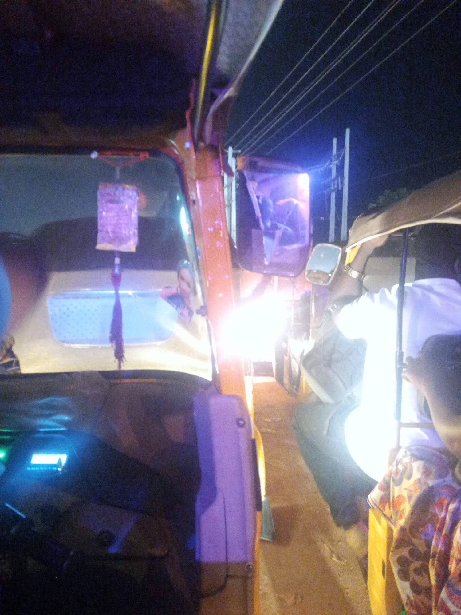 The image captures a nighttime scene in Ado, Nigeria, from the interior perspective of a vehicle, likely an auto-rickshaw or similar three-wheeled transport.
In the upper-center, a rectangular, light-reflective object, appearing purple under the ambient light, hangs from an unseen attachment point above the windshield. A dark, braided tassel extends downwards from its base. Below and behind this object, through what appears to be a rear window or partition, the upper body and face of a person are partially visible, illuminated by a soft light. This window has a faint, perforated pattern.
On the vehicle's interior right, a structural pillar or frame is present, displaying an orange-yellow hue under the dominant lighting conditions. The lower-left portion of the image shows an illuminated digital display panel with blue-green text, consistent with a vehicle's dashboard or audio system.
The exterior scene is characterized by intense artificial illumination. A very bright, white light source, possibly headlights from an oncoming vehicle, creates significant glare on the right side, overexposing a large portion of the image. This light is reflected in the vehicle's right-side mirror, which shows a distorted, bright reflection.
To the right of the vehicle, a person is visible wearing a white collared shirt and dark trousers. A gold-colored watch is worn on their left wrist. Their arm is raised and positioned towards the upper right of the frame. Below this individual, another person's leg, clad in blue jeans, is visible. Adjacent to them, a multi-colored fabric with floral patterns in orange, white, and dark tones is present. The ground beneath the individuals and vehicle appears to be an unpaved, light brown surface.
In the upper-right background, against a dark night sky, two vertical white utility poles are discernible, supporting multiple parallel power lines. A distinct blue light source emanates from an area below these power lines.