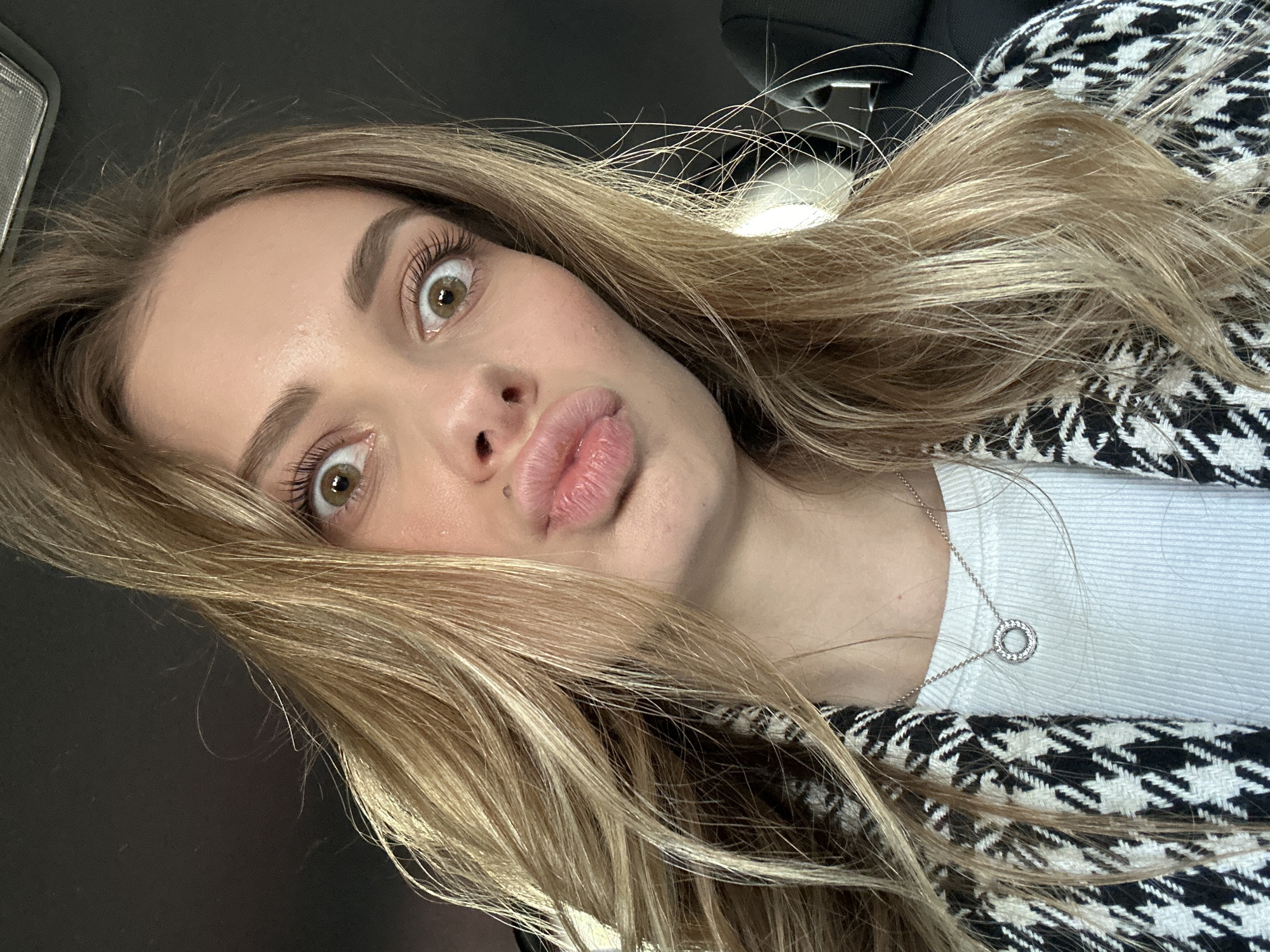 A woman with blonde hair, wearing a white shirt and a black and white houndstooth pattern jacket, is pictured indoors, likely in a car. The woman is making a "duck face" expression with her lips. She is also wearing a silver necklace with a circular pendant. The interior of the car is visible, with a black dashboard and overhead console. There is no indication of Warsaw, Poland.