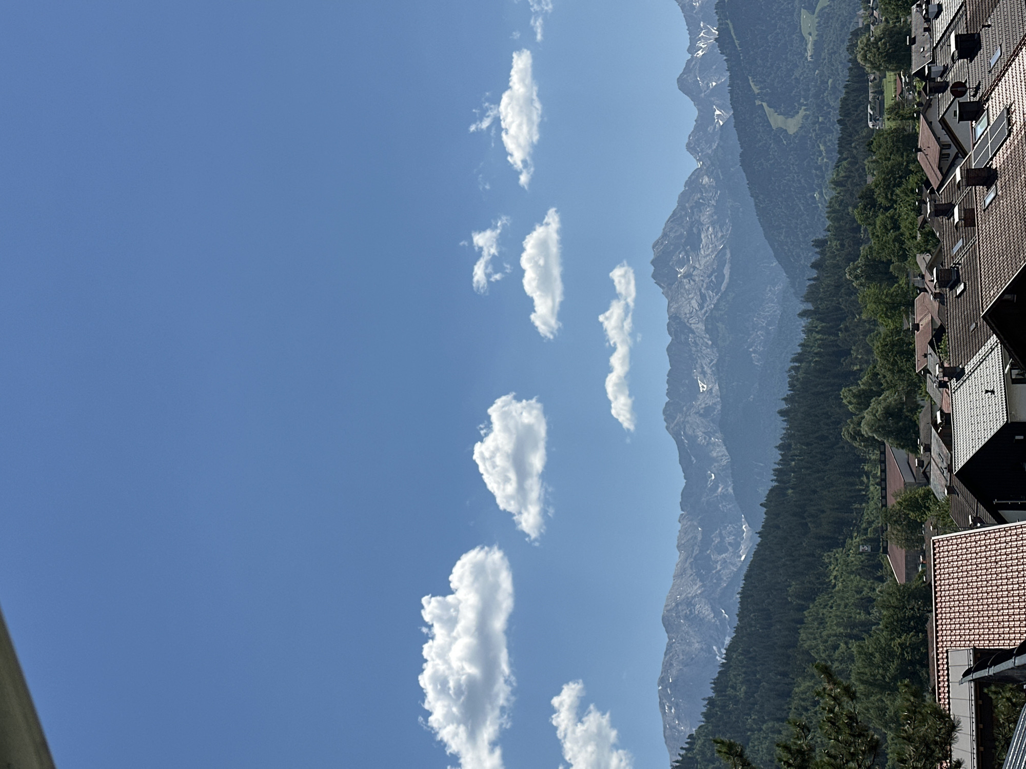 This image captures a scenic view of the Bavarian Alps in Garmisch-Partenkirchen, Germany. The scene is dominated by a clear blue sky with scattered fluffy white clouds. To the right, majestic mountains rise, their peaks partially dusted with snow, indicating a cooler higher altitude even on a presumably warm day. Below the mountains, a dense, lush green forest blankets the landscape, giving way to a village with buildings featuring red-tiled roofs. The architecture suggests a European setting, likely alpine. The perspective is from a high vantage point, possibly a balcony or rooftop, looking out over the town and towards the natural grandeur of the mountains. The time of day appears to be daytime, with bright sunlight casting clear shadows, typical of a sunny afternoon. No people or specific activities are visible, focusing the attention on the interplay of natural elements and human settlement. The overall impression is one of tranquility and natural beauty.