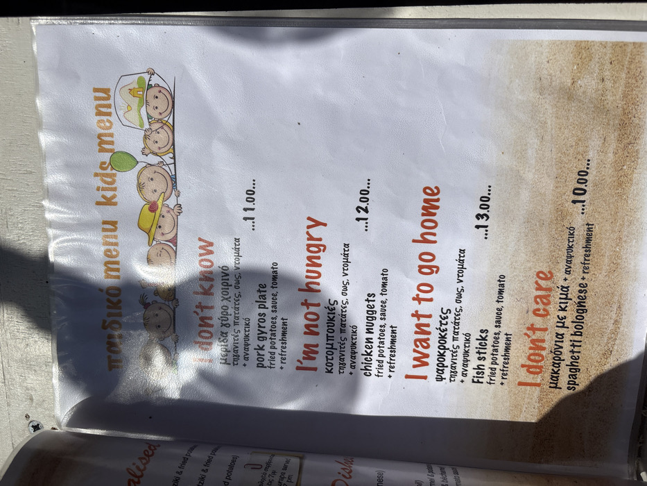 The image displays a laminated children's menu from a restaurant located in the city of Saint Arsenius, Greece. The menu's top section features the title "παιδικό menu kids menu" in orange and green text, along with an illustration of five cartoon children's faces peeking over a horizontal line. One child wears a yellow hat, another holds a green balloon, and a third wears a hat depicting a building with mountains.
Four menu items are presented with humorous English titles in red font: "I don't know" (pork gyros plate), "I'm not hungry" (chicken nuggets), "I want to go home" (fish sticks), and "I don't care" (spaghetti bolognese). Each item includes supplementary details in black text: the first three list "fried potatoes, sauce, tomato + refreshment," while the last lists "+ refreshment." Corresponding prices of 11.00, 12.00, 13.00, and 10.00 are listed next to each item. The bottom portion of the menu features a printed sandy texture as a background element.
The menu is positioned outdoors under direct sunlight, which creates significant glare across its laminated surface, particularly in the central and upper-right areas. Dark, angular shadows, likely from an overhead object or person, are cast prominently across the menu, obscuring parts of the text and illustrations on the left side. The menu exhibits signs of use, including minor creases and smudges. A partial view of an adjacent, bound menu page is visible along the left edge of the frame.