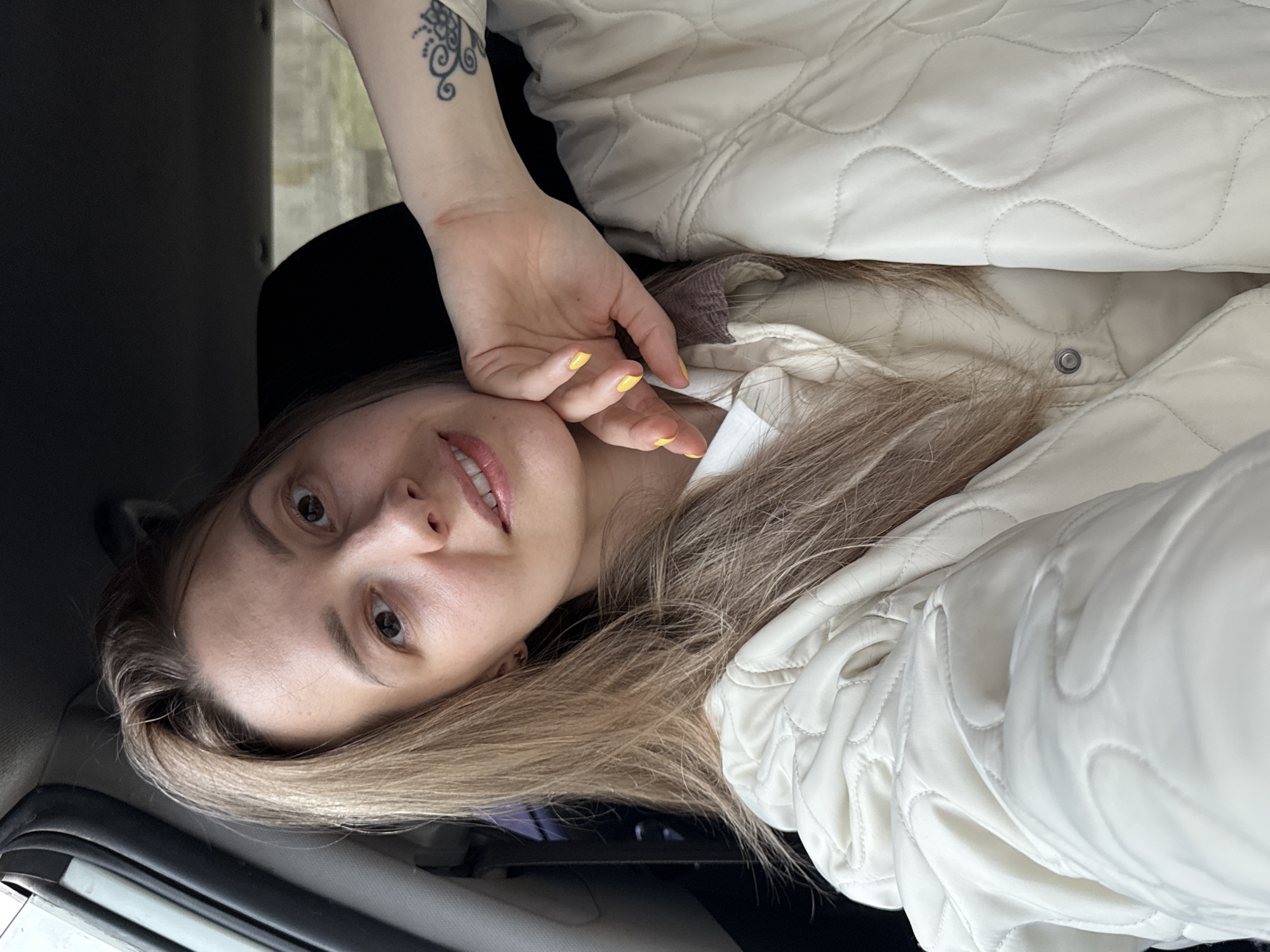 A young woman with light brown hair and yellow nail polish is in a car. She is wearing a white quilted jacket. Her left hand with a tattoo is resting on her face. The scene is likely in Chaikovsky, Russia, based on the request. The inside of the car is visible around her. The environment is partially visible through the window behind her arm.