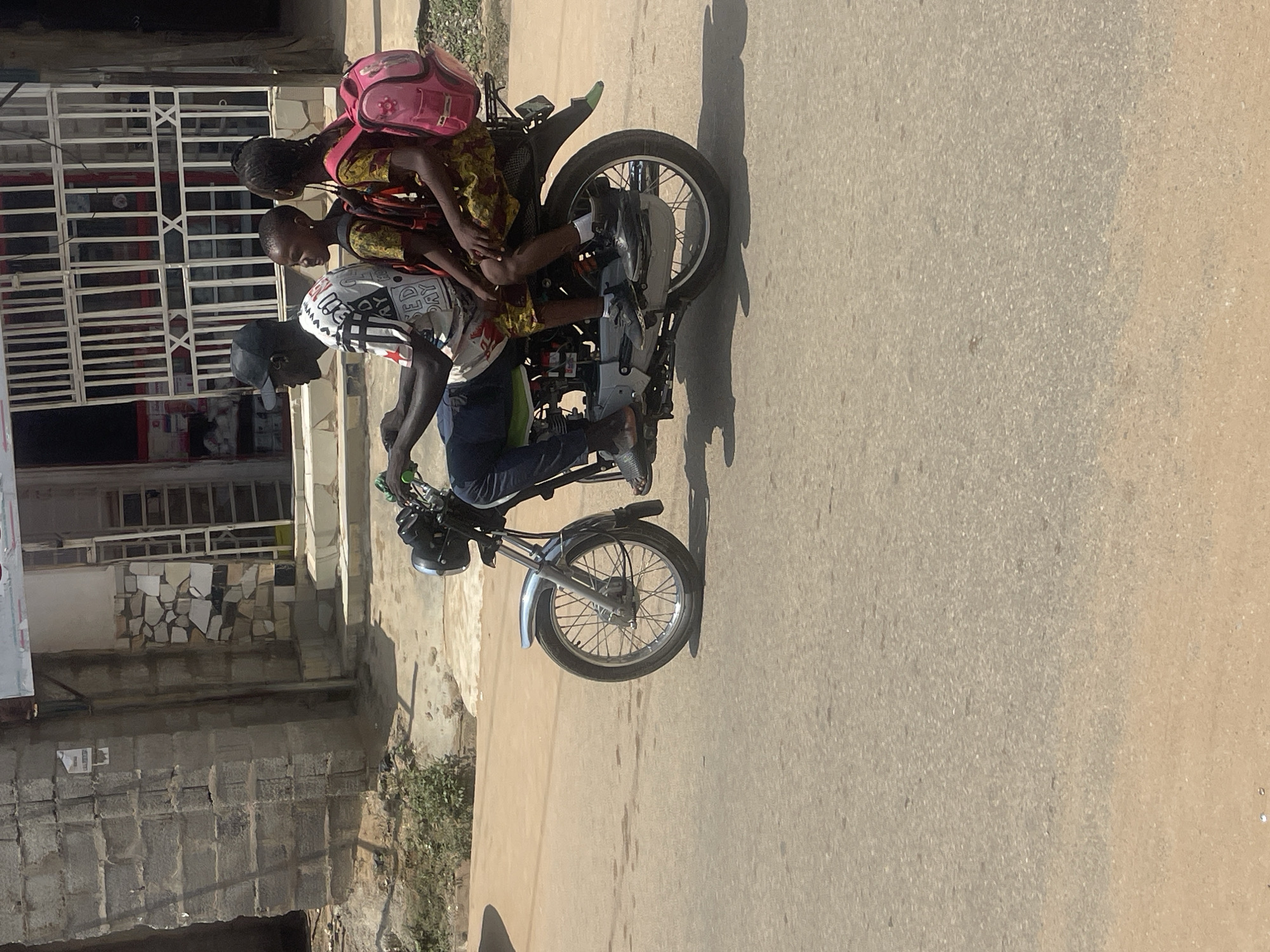 A man wearing a black baseball cap, a white t-shirt with black and red lettering, and dark blue jeans is riding a motorcycle. He is followed by two young girls. The girl in the middle wears a yellow and brown patterned dress and has a backpack on her shoulders. The girl behind her wears a similar dress and has a bright pink backpack. They are on a paved road, with a building and vegetation visible in the background. The image appears to be taken in Abuja, Nigeria, given the context.