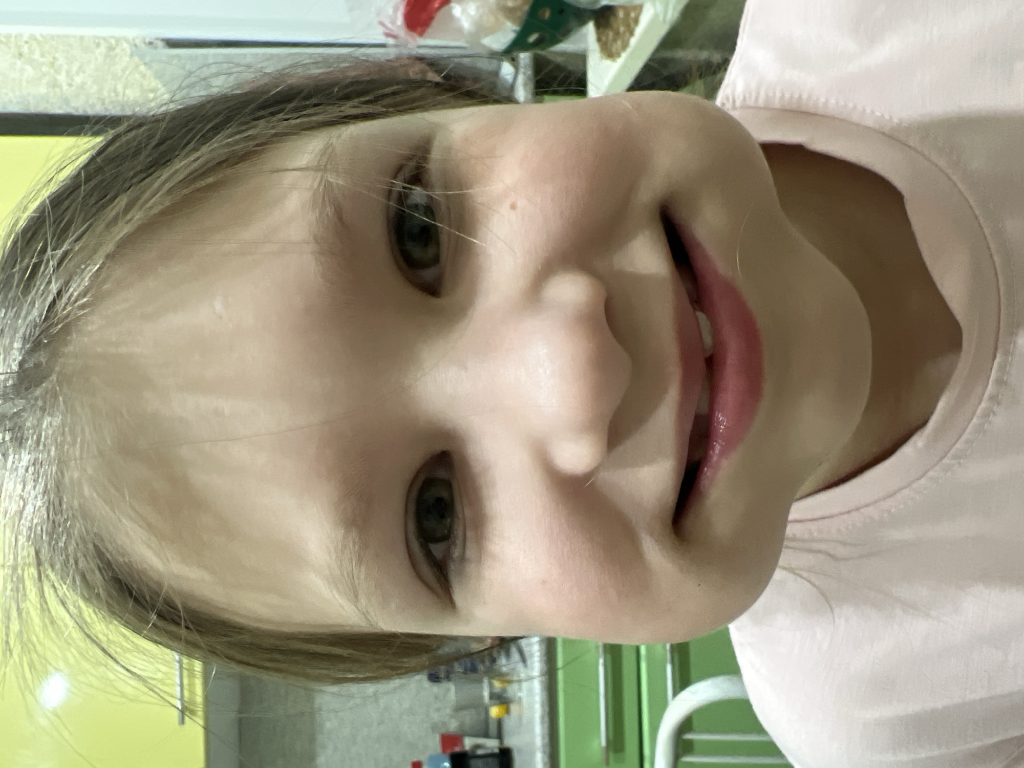 A young child with light brown hair and dark eyes is depicted in a close-up portrait, smiling broadly with visibly puffed cheeks, facing directly towards the camera. The child wears a light pink t-shirt. The scene is located in a kitchen in Chaikovsky, Russia. The background features light yellow overhead cabinetry and light green lower cabinets with silver handles. A white chair is partially visible on the lower left. Various blurry kitchen items, including bottles and a green bowl with contents, are partially visible on a counter surface to the right.