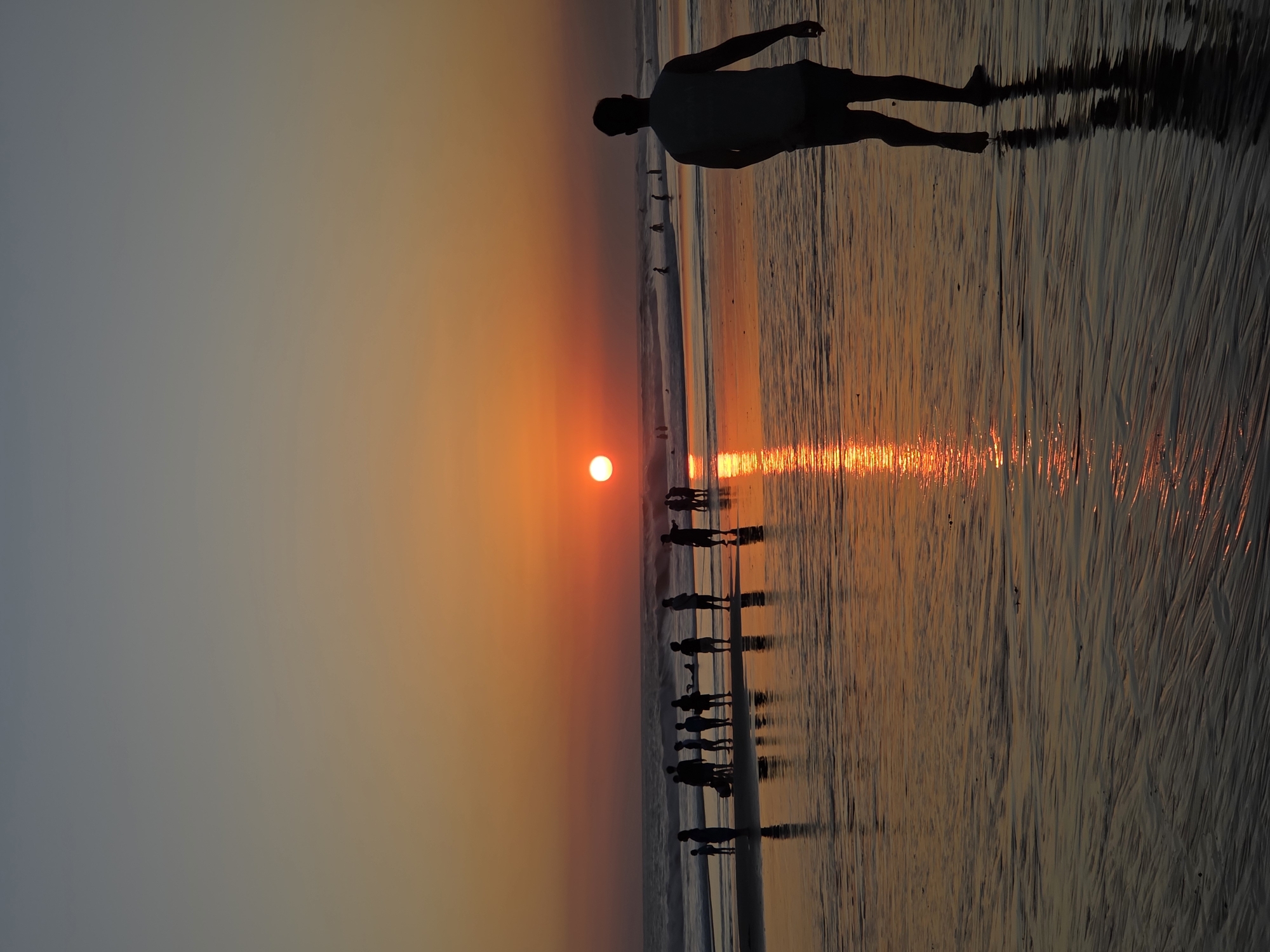 A man is walking on a beach at sunset. The sun is a bright orange orb low on the horizon, casting a warm glow across the sky and water. The wet sand reflects the colors of the sunset, creating a shimmering surface. In the distance, other people can be seen on the beach and in the water, silhouetted against the bright sky. The scene appears to be in North Kuta, Indonesia, a popular tourist destination known for its beaches and sunsets. The image captures a serene and peaceful moment as the day comes to a close.