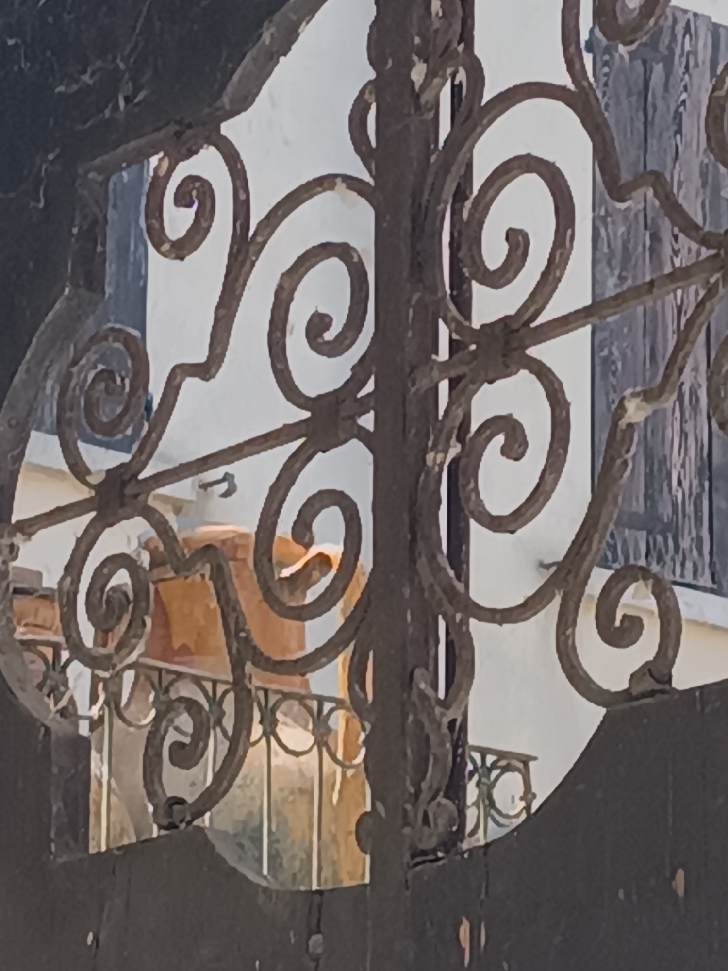 A close-up photograph shows a decorative wrought-iron gate or fence in Mezzana Sant'Antonio, Italy. The ironwork features elaborate scroll designs and appears aged, with paint flaking off in some areas. The background visible through the ironwork includes a white building with a closed blue shutter and a balcony with a black railing. A tan-colored object is partially visible on the balcony. The overall composition is centered on the intricate details of the ironwork, partially obscuring the details of the building behind it.