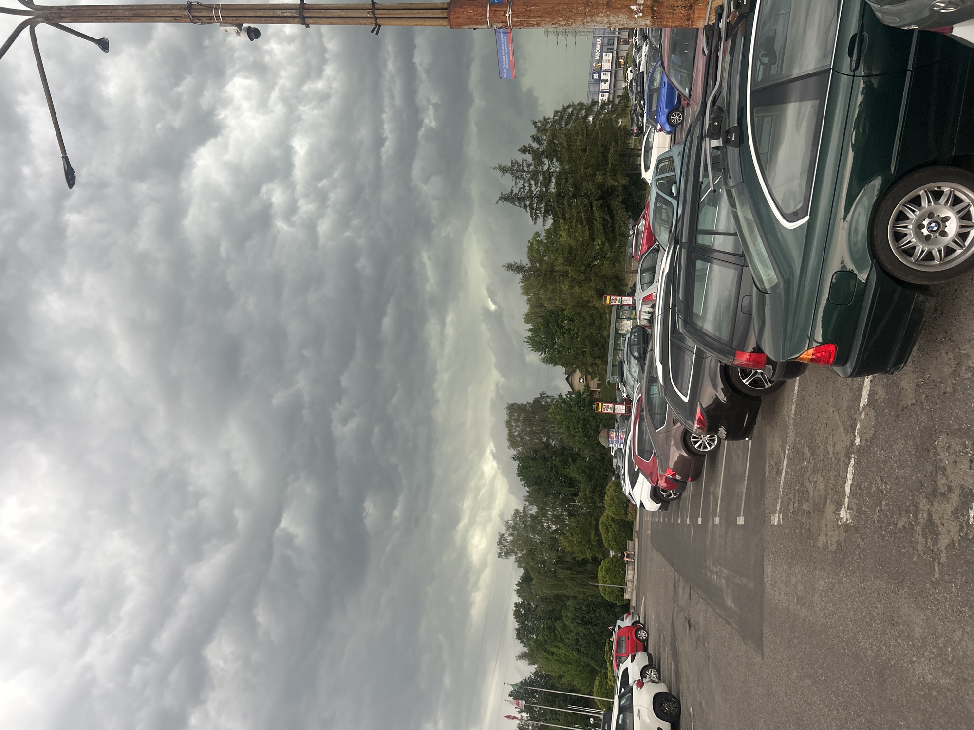The image shows a parking lot filled with cars under a cloudy sky. The cars are parked in rows on an asphalt surface marked with white lines. In the foreground, a dark green BMW station wagon is visible, featuring a roof rack and silver alloy wheels. Several other cars, including a blue hatchback, a red hatchback, and white cars, are also present.
Behind the cars, a line of trees with lush green foliage separates the parking lot from a few buildings. A sign for "MRÓWKA" is visible on one of the buildings, suggesting it might be a retail store. The sky is filled with dramatic, dark gray clouds, indicating overcast weather, possibly before or during a storm. The overall lighting suggests it is daytime, though the cloud cover makes it appear dim.
The image is oriented vertically, with the sky occupying the left half and the cars and parking lot on the right. A wooden structure with what appears to be lighting fixtures runs along the top edge of the image. There are no people clearly visible in the frame, and the overall mood is somber due to the weather. The location is identified as Jastrzębie-Zdrój, Poland.