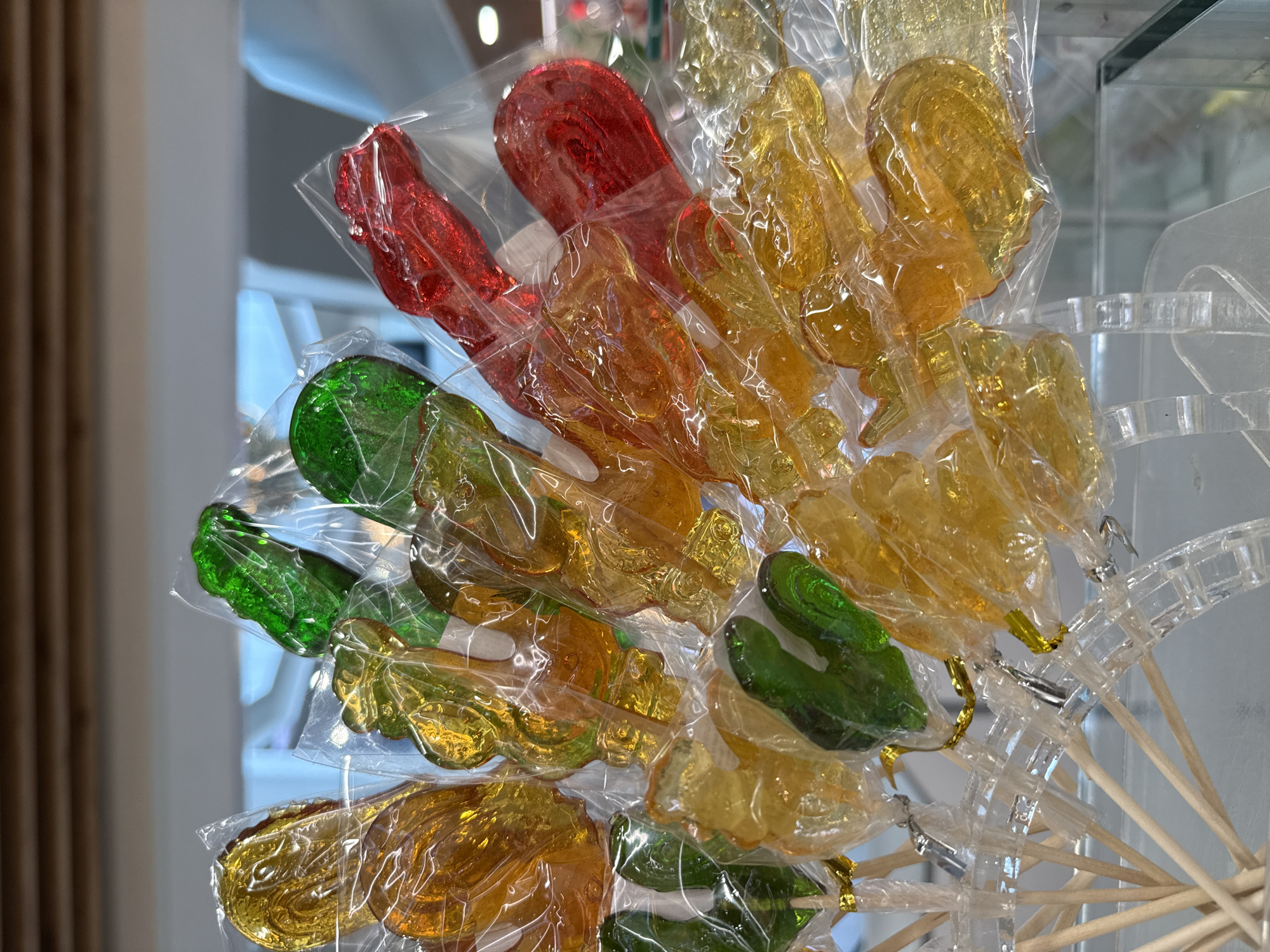 The image shows a close-up view of a display of colorful, animal-shaped lollipops. The lollipops are individually wrapped in clear plastic and are arranged in a fan-like pattern, with sticks inserted into a clear plastic holder at the bottom. The lollipops are a variety of colors, including red, orange, yellow, and green, and appear to be made of hard candy. Some of the lollipops are shaped like fish, while others have more abstract, swirling designs. The background is blurred but appears to be a retail environment with shelves and lighting. The image does not provide any information about the specific location in Izhevsk, Russia.