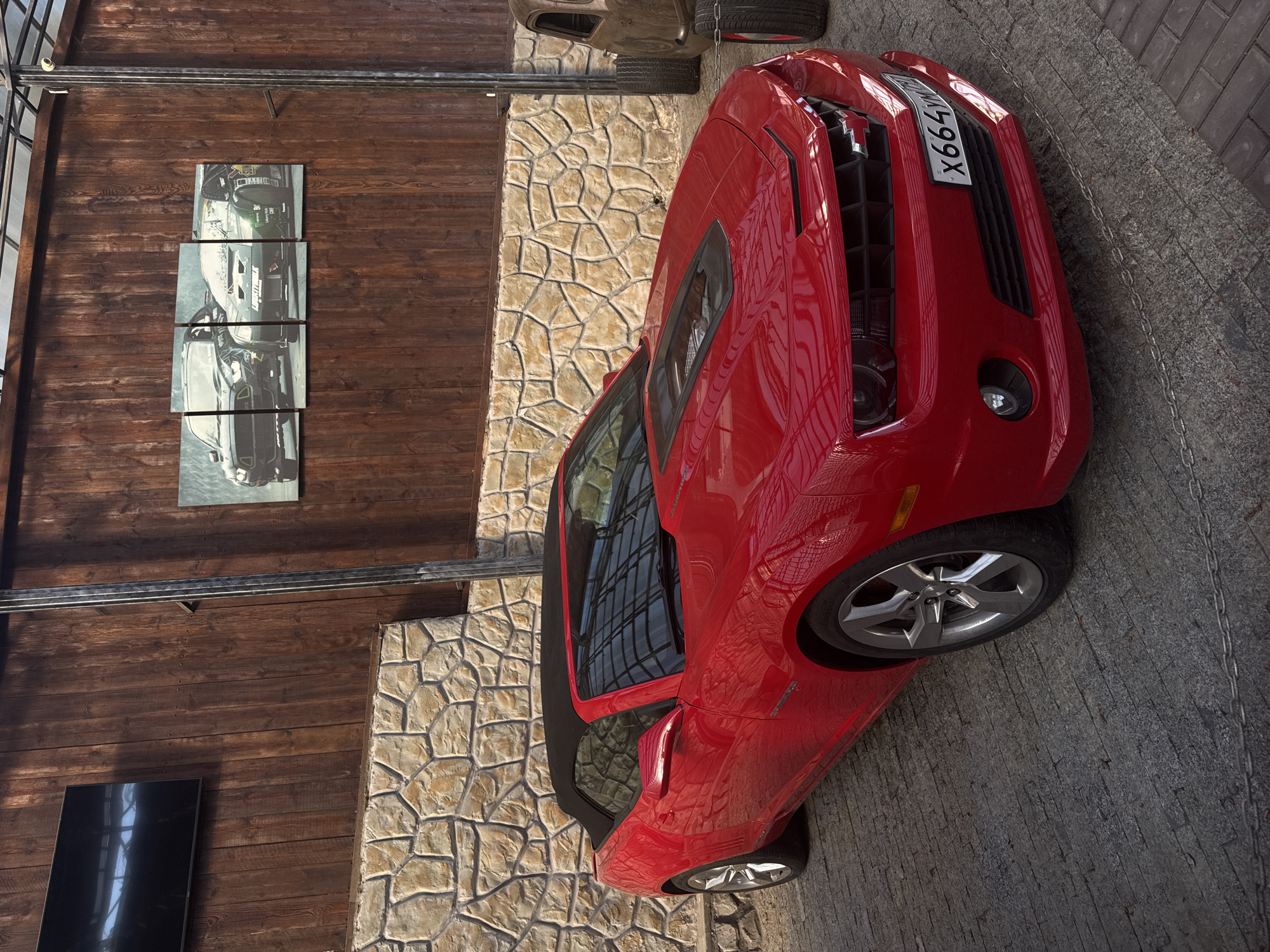 The image shows a red Chevrolet Camaro convertible parked on a paved surface. The license plate reads "X664YX". Behind the car, there is a stone wall and a wooden wall with a series of four pictures and a rectangular black object mounted on it. Another, partially visible vehicle can be seen above the Camaro. The location is likely in Ялта, Ukraine based on the license plate. The scene appears to be an outdoor parking area.