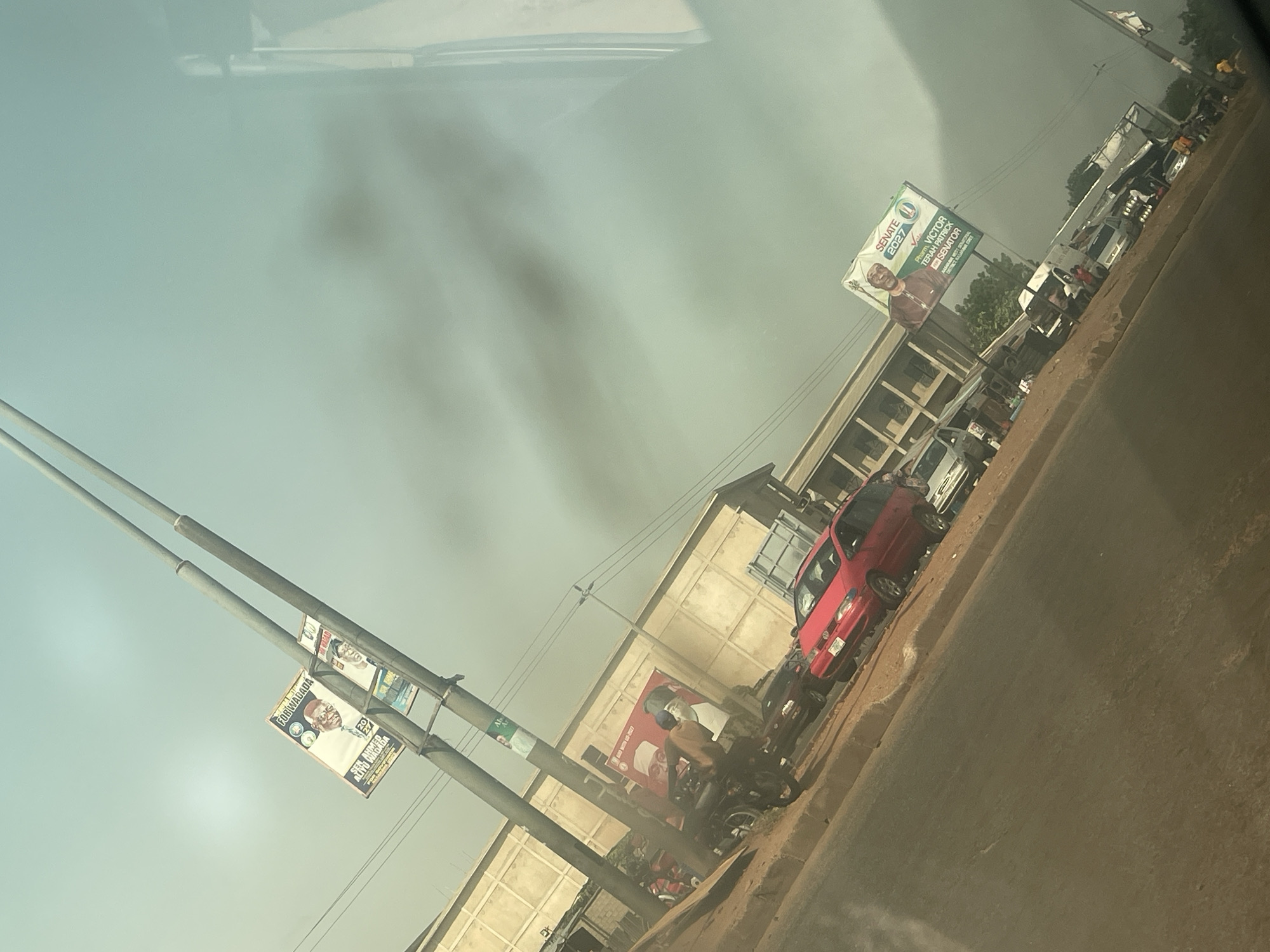 This image, taken from inside a vehicle and oriented diagonally, depicts a hazy daytime street scene in Keffi, Nigeria. The foreground shows a paved road and a dirt shoulder where several vehicles, including a red car and multiple motorcycles, are parked or moving slowly. One individual is visible on a motorcycle, dressed in light-colored attire.
Along the roadside, various buildings with light-colored or unpainted facades line the street. The sky above is bright but appears notably hazy or dusty, suggesting dry weather conditions.
Prominent in the scene are several utility poles and billboards adorned with political campaign posters. One large billboard clearly displays "SENATE 2027" and features "Pharm. VICTOR TERAH PATRICK" as a "SENATOR" candidate. On a nearby utility pole, additional posters are visible with text such as "FOR WADADA" and "ALFAO WAMEDA," also indicating "20 27," likely referring to the same upcoming election year.
The image captures a bustling street environment characterized by local architecture, vehicular traffic, and prevalent political campaigning for the 2027 elections.