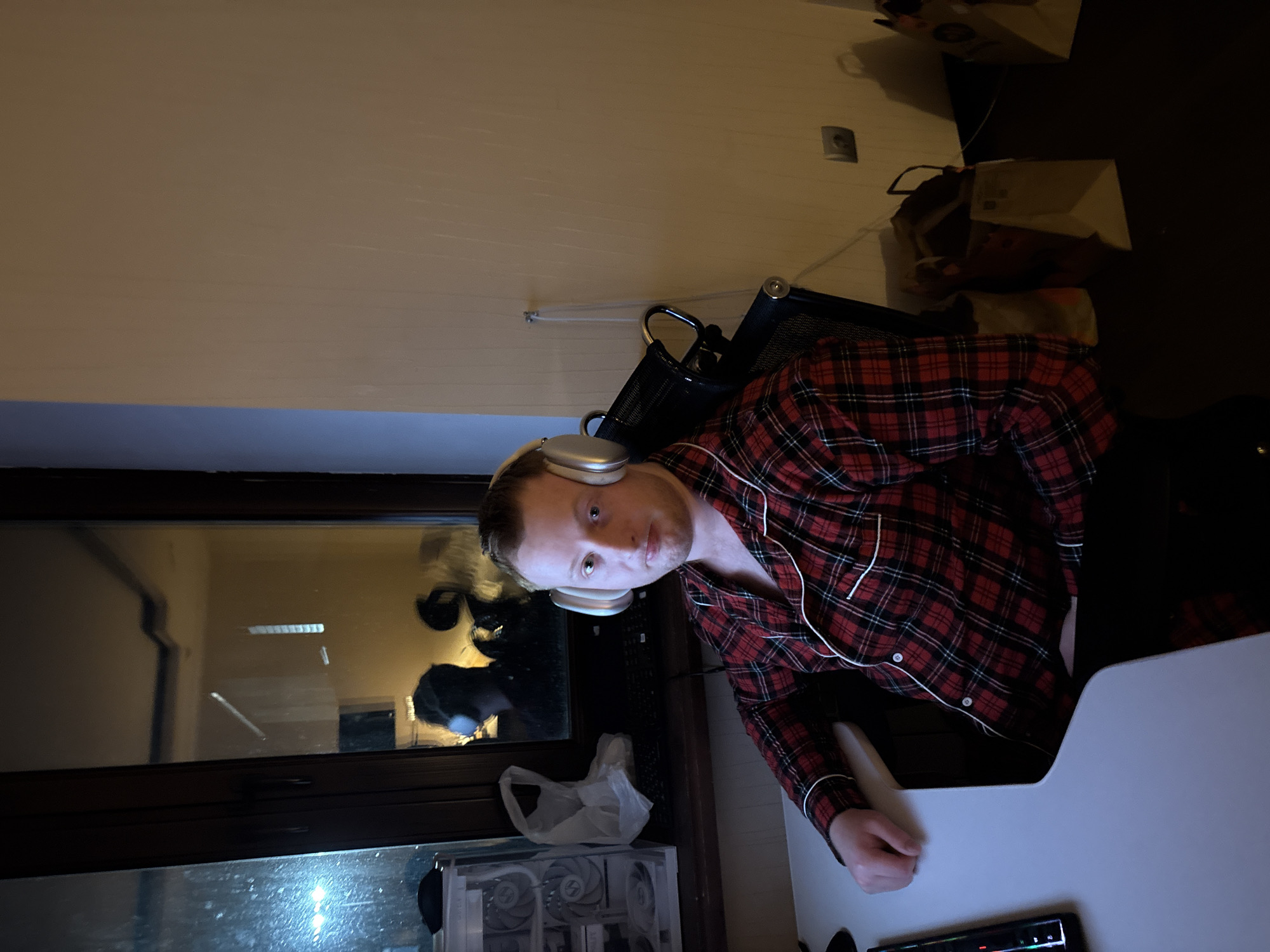 A man wearing headphones and a red plaid shirt sits at a desk. A window is visible with a reflection of two people. The desk includes a computer and peripherals. The location details (Ореховка, Russia) cannot be determined from the visual information.
