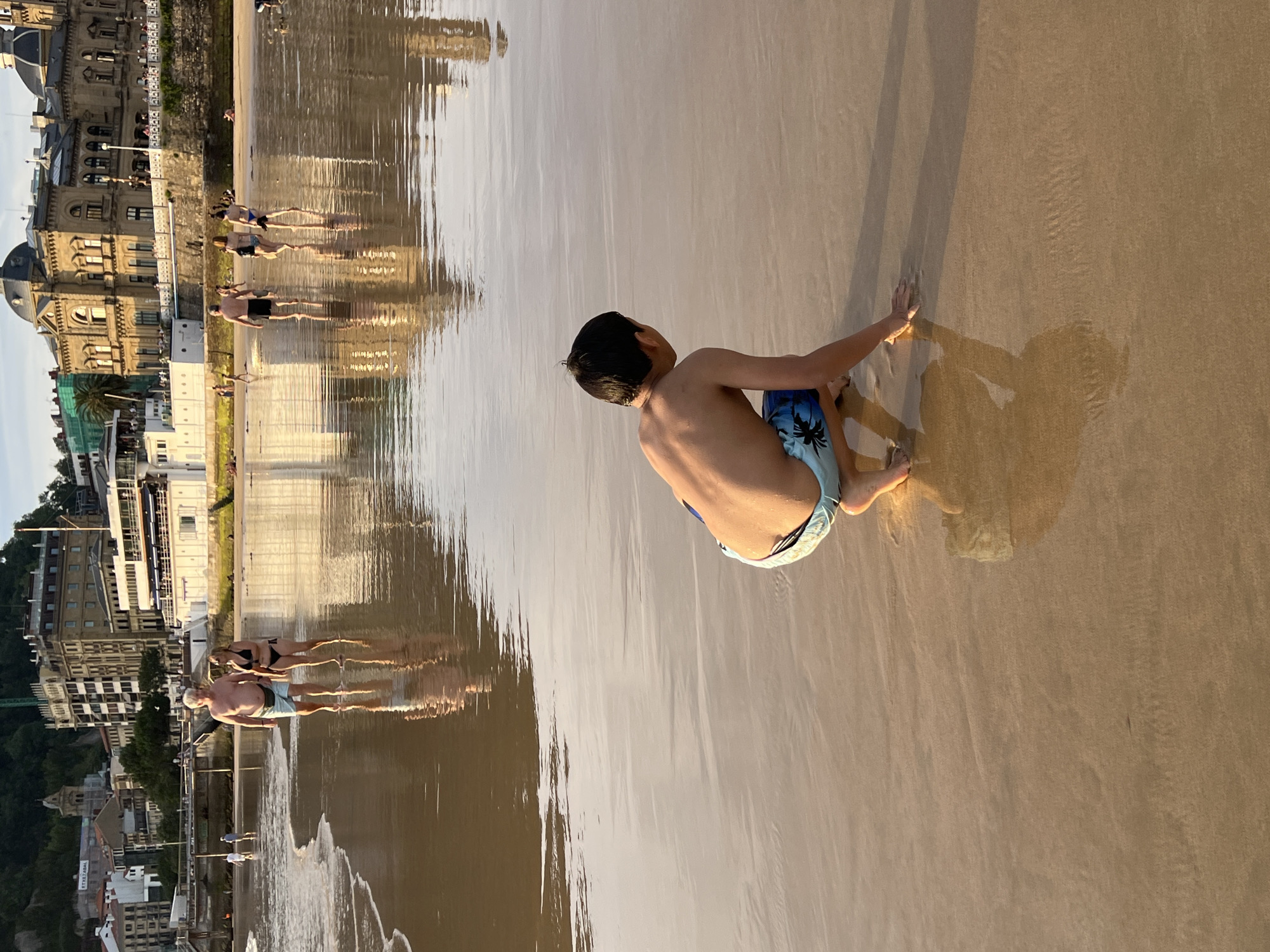 This is a vertically oriented, wide-angle photograph taken on a sandy beach in San Sebastián, Spain. The image features several people scattered across the beach and shallow water, with buildings and lush green hills visible in the background. A young boy in blue patterned swim trunks is the most prominent subject, sitting with his back to the camera on the wet sand. He is leaning forward, with his hands resting on the sand as if preparing to push himself up. His shadow, elongated by the low angle of the sun, stretches out before him.
Further down the beach, a group of people are wading in the shallow water, their reflections mirrored on the smooth, wet sand. They appear to be enjoying a leisurely day at the beach. The lighting suggests it is either early morning or late afternoon, with the sun casting long shadows and creating a warm, golden hue across the scene. The water is calm and reflective, adding to the serene atmosphere. The architectural style of the buildings in the background, with their ornate facades and balconies, hints at a European coastal city. There is no visible text in the image.