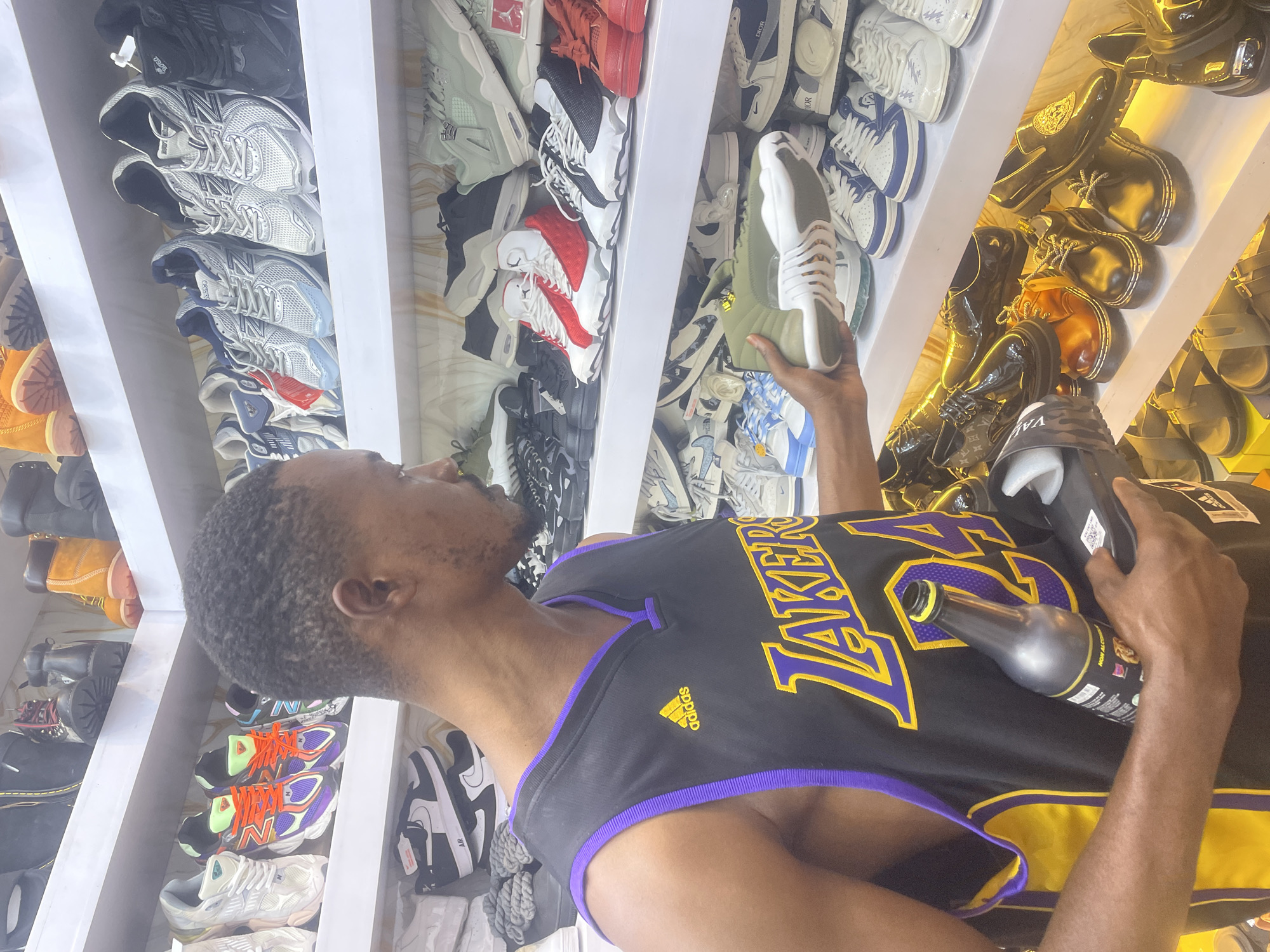 A man in a black and yellow Lakers jersey is in a shop with rows of sneakers and shoes on display shelves. He's holding a sneaker in one hand and a bottle in the other. The location is likely in the city of Jos, Nigeria, given the request for that detail.