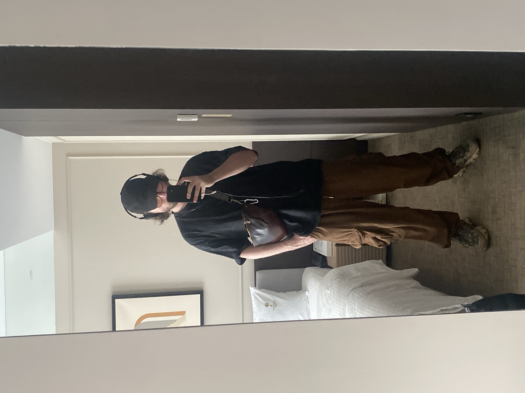 The image depicts a person taking a selfie in a room. The individual is wearing a black t-shirt, brown pants, a black cap, and headphones. They have a brown bag slung across their body. The room includes a bed with white linens, a painting hanging on the wall, and a mirror reflecting the scene. The location in the city of Elino, Russia, cannot be determined from the provided image.