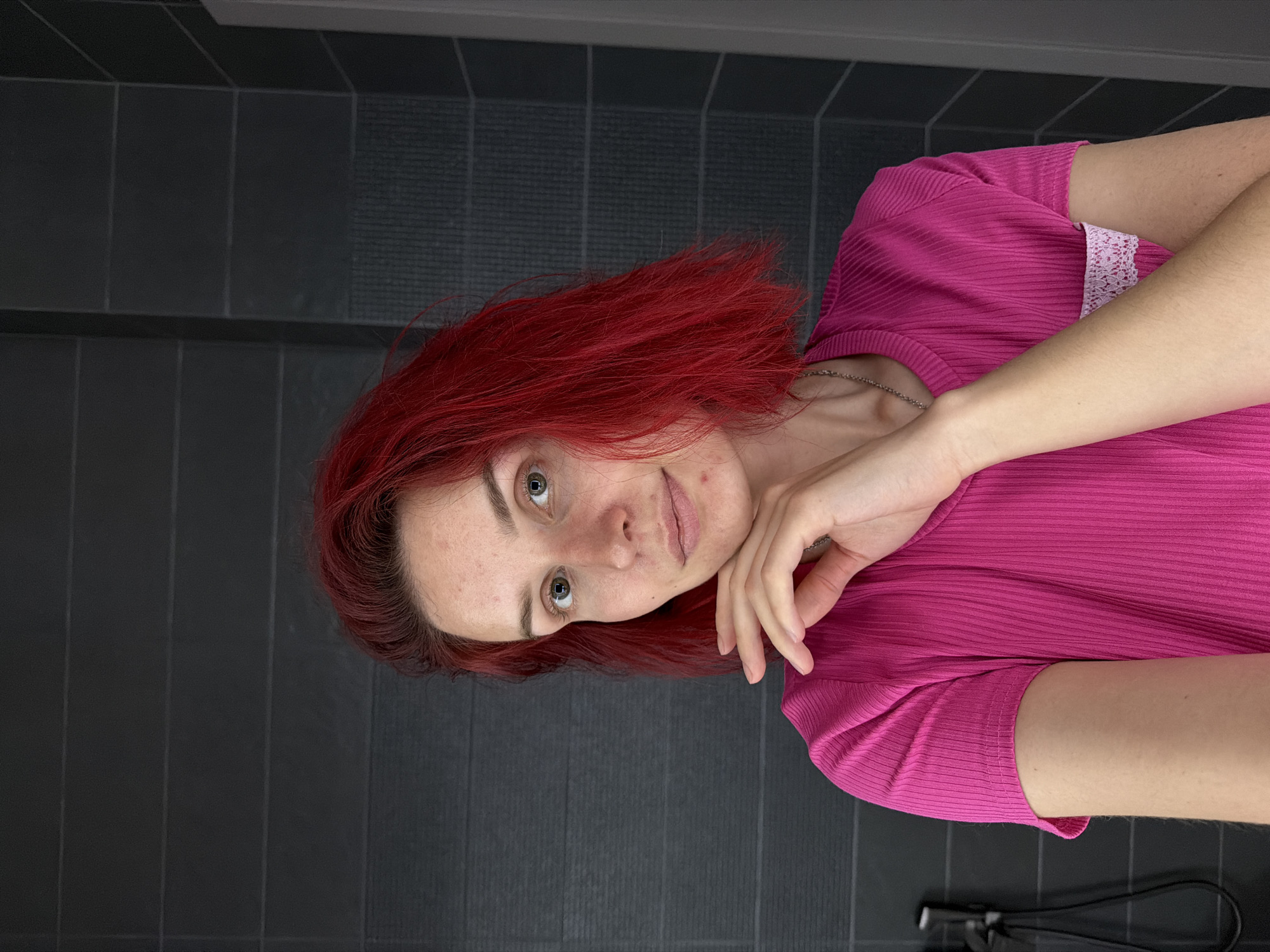 A woman with red hair and green eyes is wearing a bright pink ribbed shirt with lace trim. She rests her chin on her hand and looks towards the camera. She is in front of a gray tiled wall. The image may have been taken in a bathroom in Chaikovsky, Russia, given the tiled wall and potential hair styling tools at the bottom of the image.