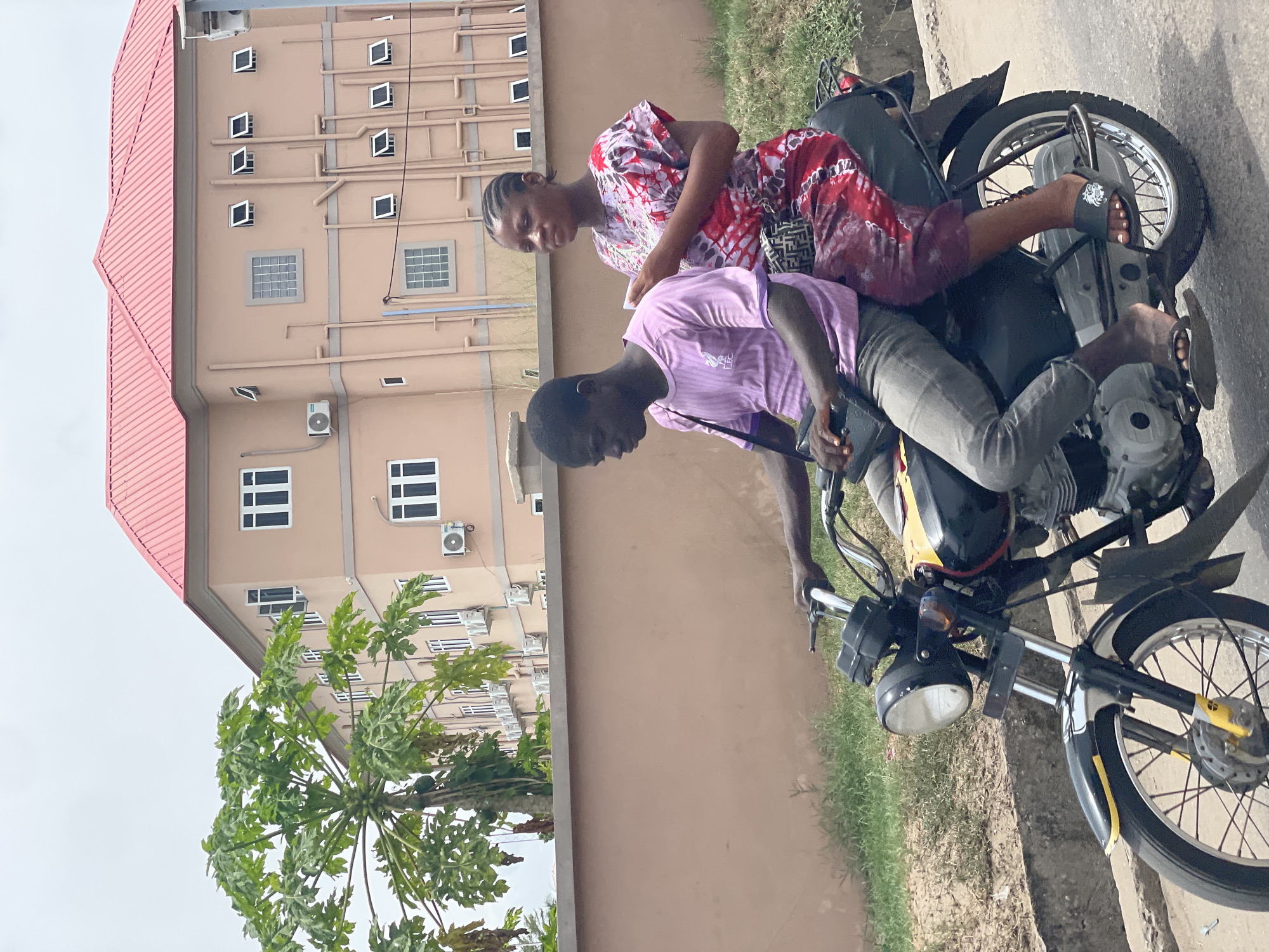 Two people are on a motorcycle. A woman in a red and white dress sits on the back. A man in a purple shirt sits in front. The motorcycle is on a paved area. Behind them is a beige building with a red roof and several windows and air conditioning units. A papaya tree is next to the building. The location is Badagry, Nigeria.