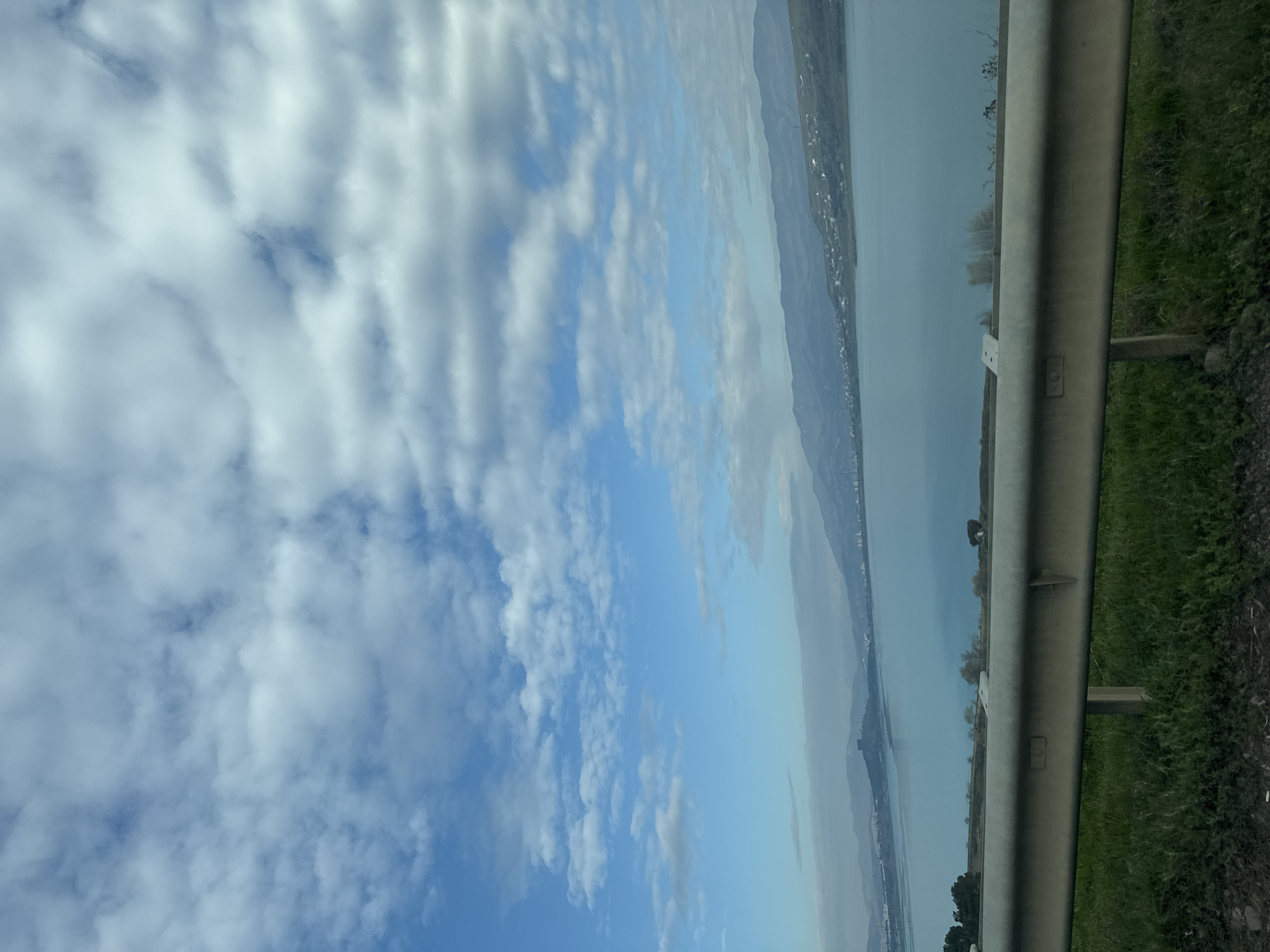 The image shows a view from what appears to be a roadside in an area near a body of water. The sky is predominantly blue with formations of white clouds. A metal guardrail runs horizontally across the right side of the frame, with green vegetation growing behind it. Beyond the guardrail and water, a stretch of land is visible. Given the proximity to water and the landscape, it is possible, but unconfirmed, that this could be a location in the vicinity of the Tbilisi Sea in Georgia, though definitive identification requires more specific details not available in the image.