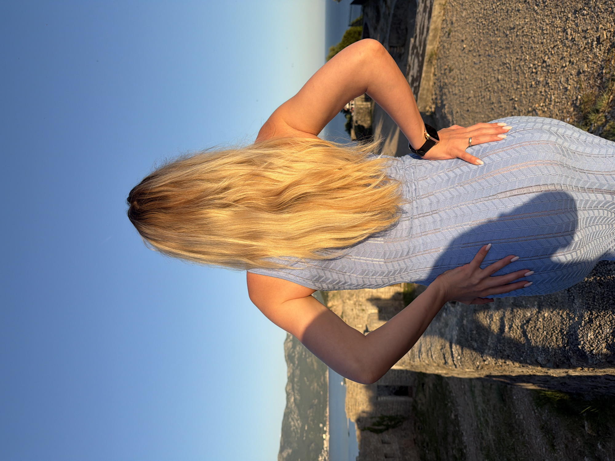 The image depicts a female individual, observed from behind, standing on a stone pathway overlooking a coastal landscape in Budva, Montenegro. She possesses long, wavy blonde hair and is dressed in a light blue, form-fitting, sleeveless knitted dress with a distinct chevron pattern. Both hands are placed on her hips; a black watch with a metallic gold bezel is present on her right wrist, and a simple gold-toned ring is visible on the ring finger of her left hand, which also displays manicured fingernails with white tips. The background features a calm body of water, likely the Adriatic Sea or Budva Bay, bordered by distant mountains under a clear blue sky. The immediate surroundings include weathered stone walls and structures, indicative of an ancient fortification or a historic area within Budva, such as the Citadel or surrounding Old Town walls. The scene is illuminated by warm, low-angle sunlight, casting elongated shadows from the individual and the stone structures onto the gravel ground.