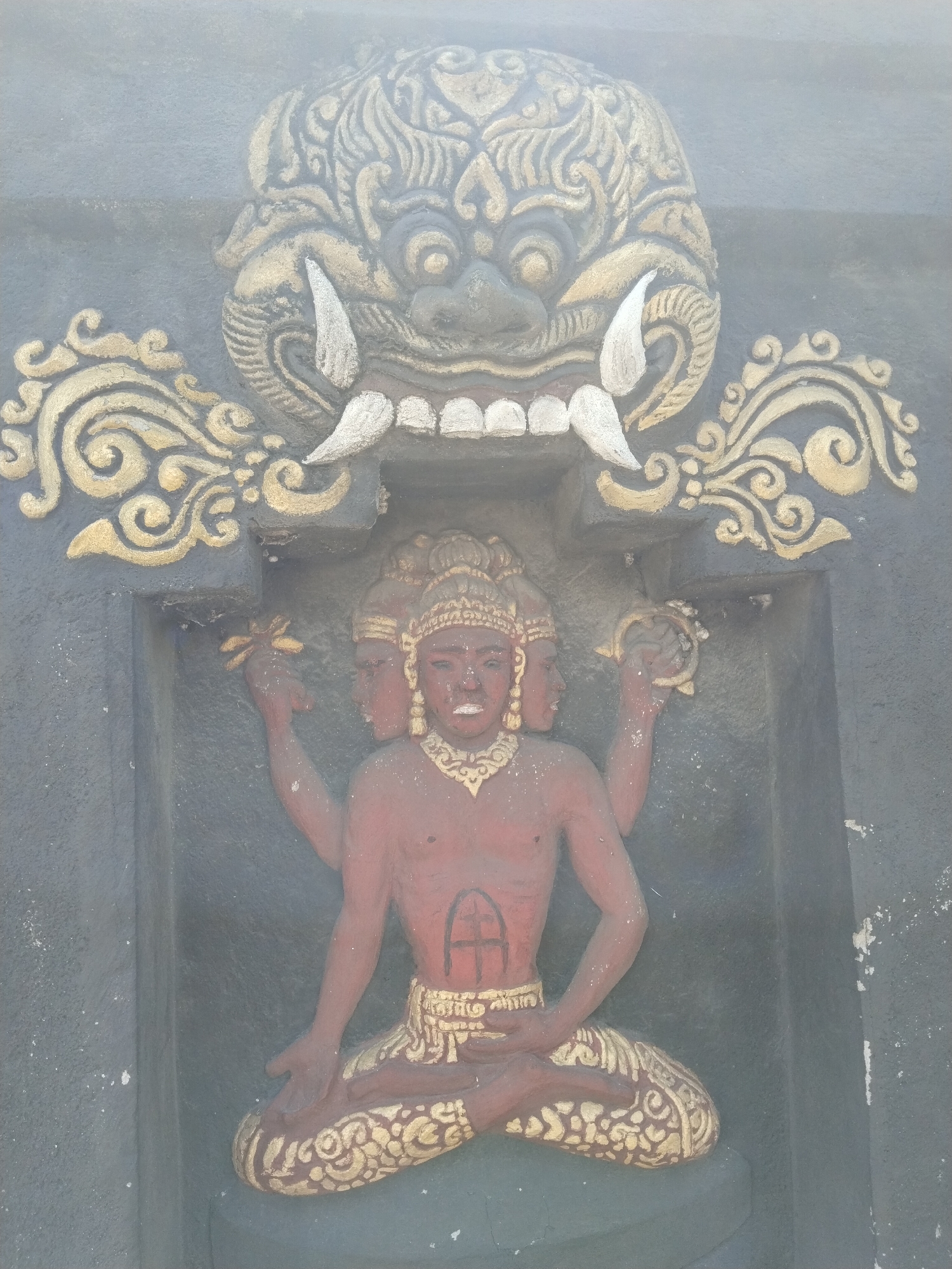The image shows a relief carving set into a grey wall. At the top is a large, ornate mask with protruding fangs. Below the mask is a four-armed, three-faced figure seated in a meditative pose. The figure is reddish-brown, adorned with gold jewelry and a patterned garment. The location is Masaran, Indonesia.