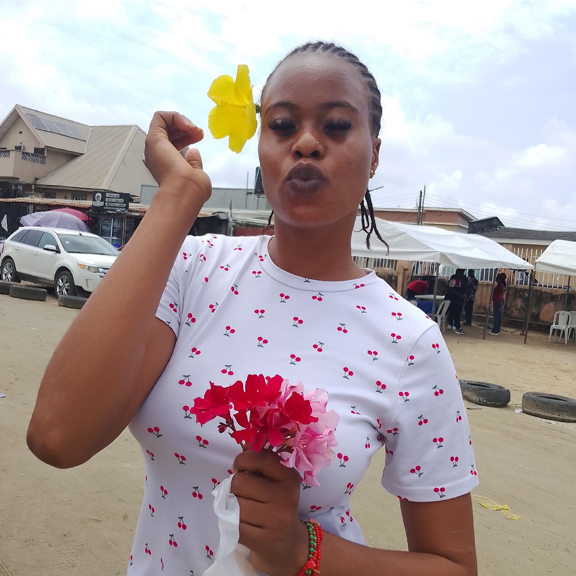 A young Black woman with braided hair is posing for a photo outdoors in what appears to be a dusty, urban environment, possibly in Ado, Nigeria. She is wearing a white t-shirt with a repeating pattern of red cherries. A single large yellow flower is tucked behind her right ear, and she holds a small bouquet of pink and red flowers in her left hand, along with a piece of white fabric. Her lips are pursed, and she has dramatic false eyelashes. In the background, there are several white tents or canopies, a white SUV, and buildings with visible solar panels. The ground is sandy or dirt. The lighting suggests it is daytime, with an overcast sky. Several people are visible in the background, some standing under the tents. There are also discarded tires on the ground.
