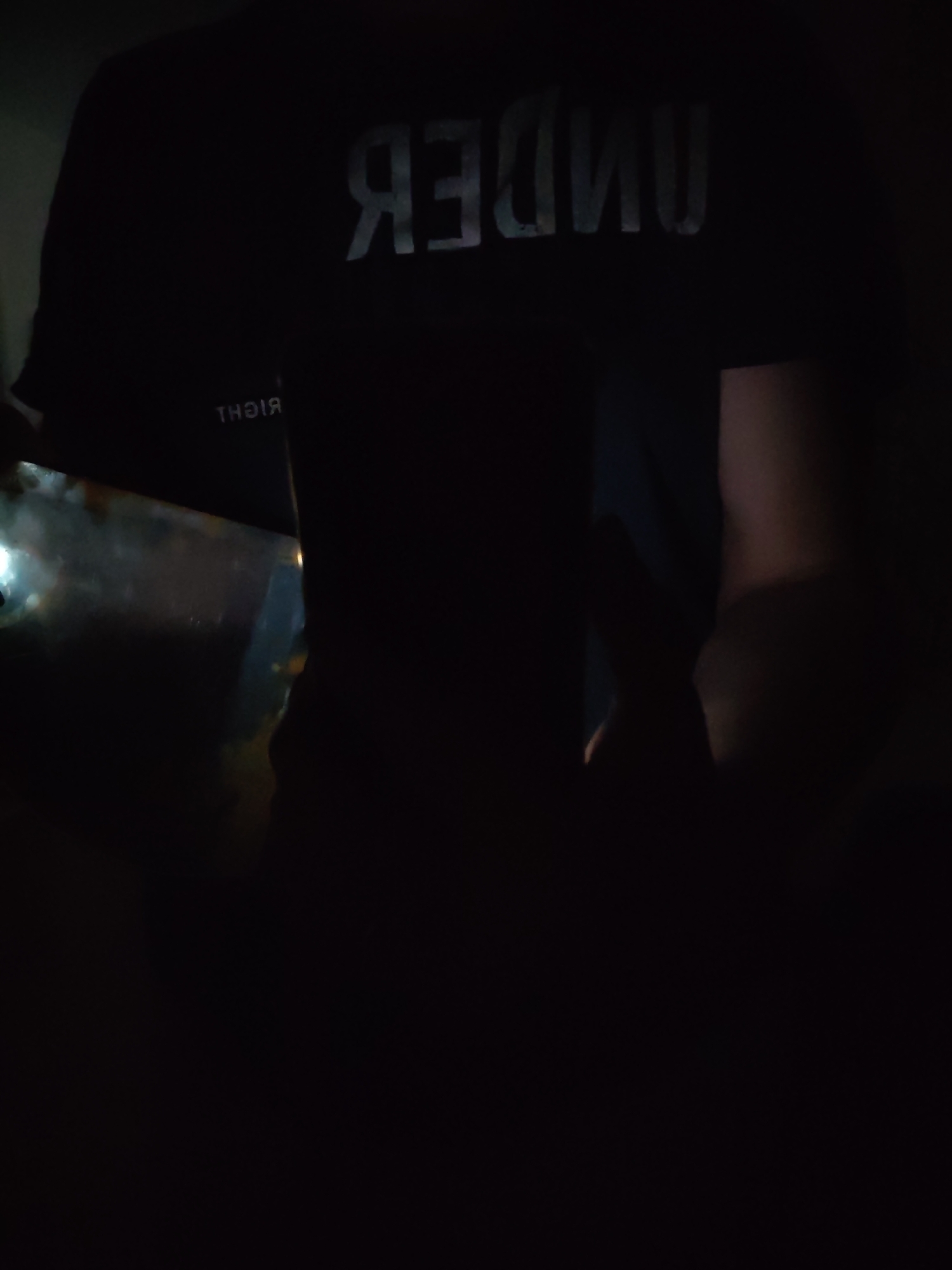 The image shows a person wearing a black shirt with the word "UNDER" printed on the back in reflective silver letters. The word "RIGHT" is printed on the sleeve. The person is standing in a dimly lit room, with some light reflecting off an object to the left. The environment is too dark to determine the location in Chaikovsky, Russia, or provide further details about the scene.