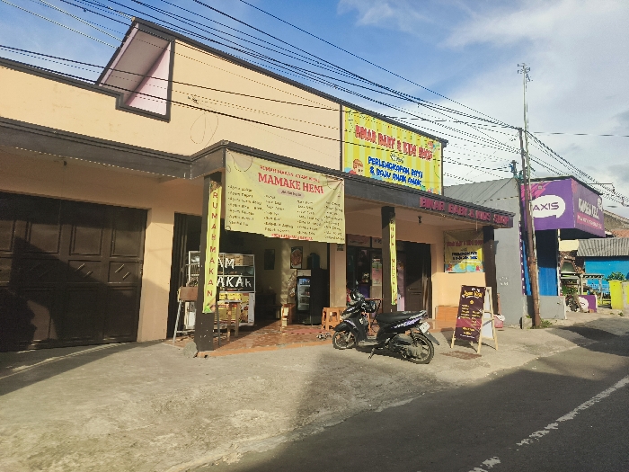 The image shows a street scene in Cikalong, Indonesia, located at latitude: -6.742, longitude: 107.442. There is a small, yellow-colored shop with a sign that says "Mamake Heati." The shop has a brown garage door to the left. In front of the shop is a parked black and silver motorcycle. The shop is on the corner and a second building with a blue sign with the word "Axis" can be seen to the right. The road in the image is paved and there are electric wires overhead. There are no people in the image. The sky is mostly blue and the sun is shining.