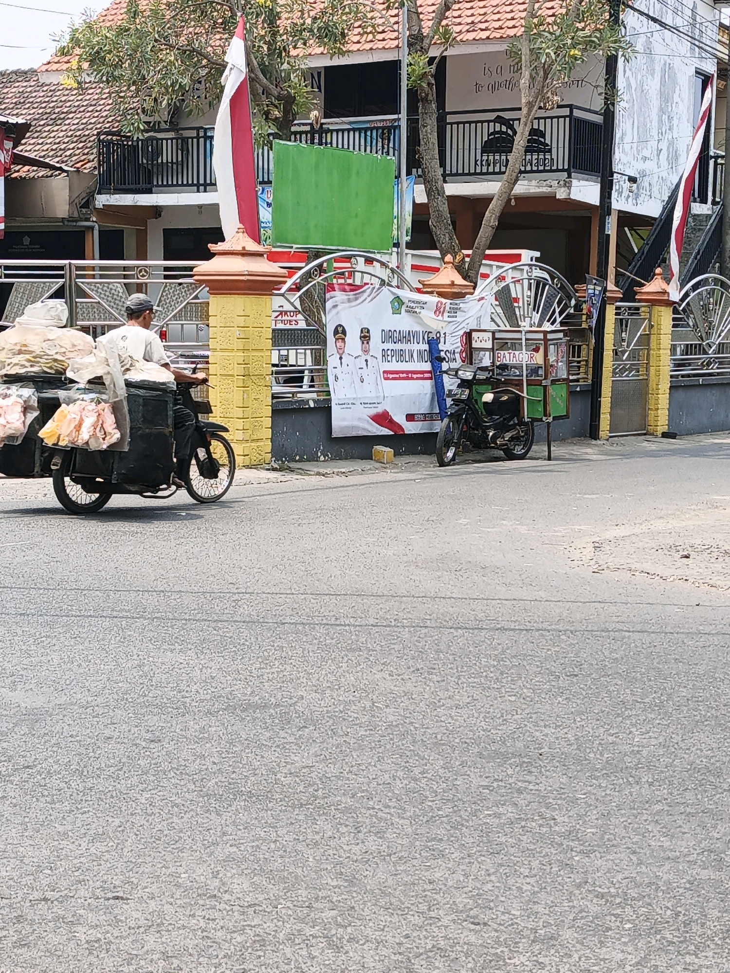 In the city of Ketegan, Indonesia, on a sunny day, a man rides a motorcycle with a sidecar full of goods. The motorcycle is dark in color, and the sidecar is also dark, appearing to be a box or container. It is loaded with large bundles of items wrapped in plastic, some appearing to be food products like vegetables or fruits. The man is wearing a light-colored shirt and a cap.
Behind the motorcycle, a building with red-tiled roofs is visible. In front of the building is a fence made of metal and yellow pillars. There are Indonesian flags displayed, one on a pole and another hanging. A banner with Indonesian text and images of men in uniform is also present. A smaller green and white cart is parked on the side, which might be a food stall. The street in the foreground is paved with asphalt. Trees with green foliage are scattered around the area. The overall scene depicts a street view with a vendor on his motorcycle, indicating a typical day in a commercial or residential area in Ketegan.